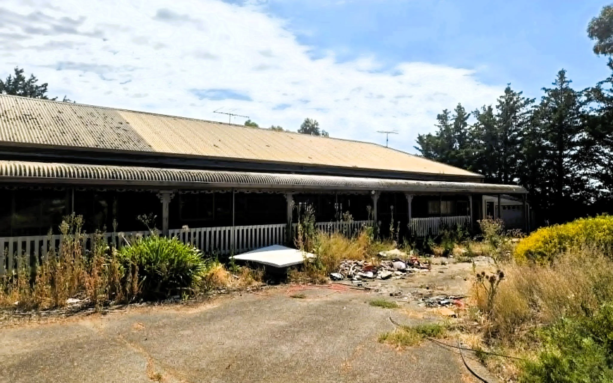 A house with a metal roof, surrounded by a garden with bushes and plants, and a white picket fence, under a partly cloudy sky.