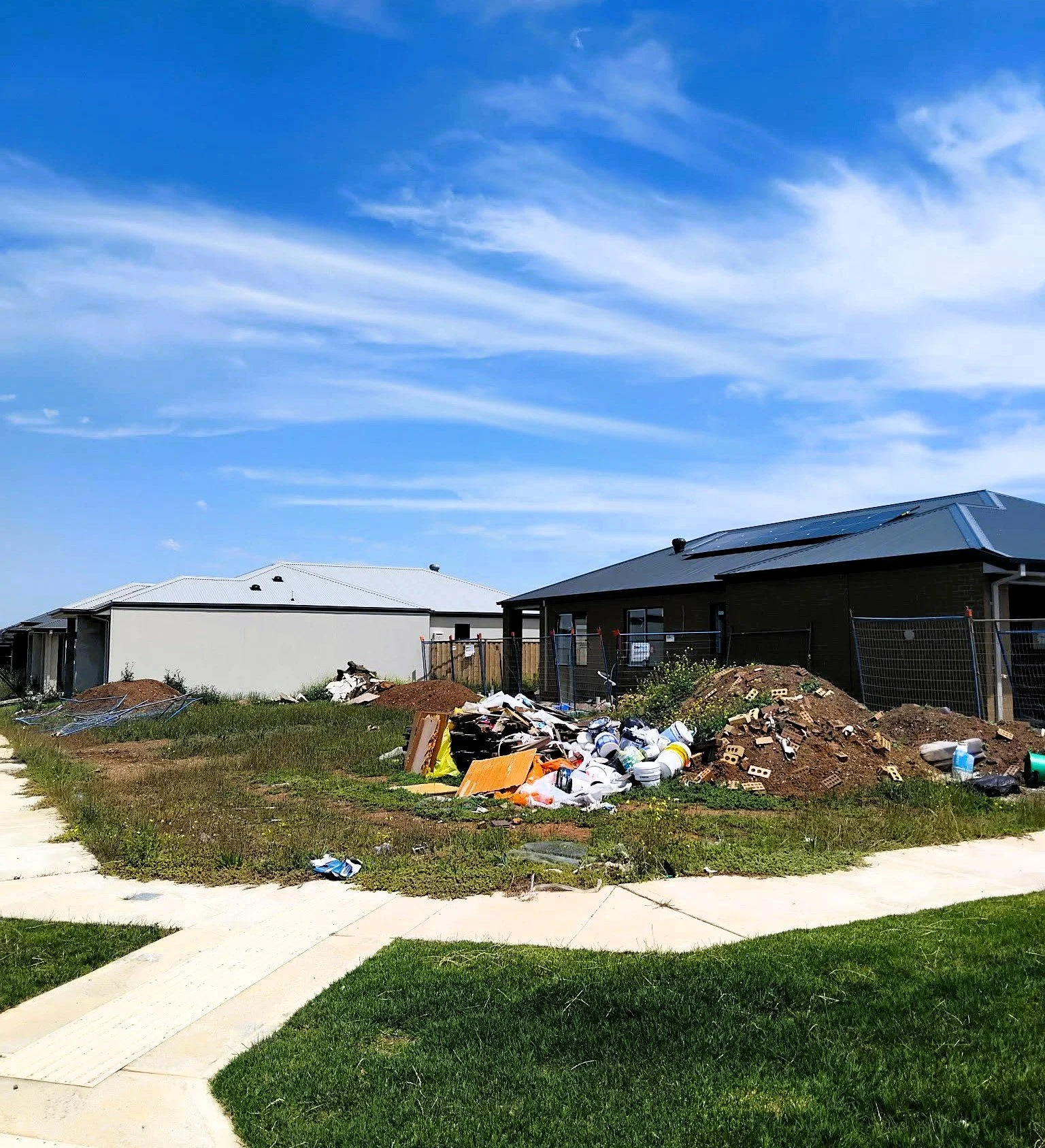 Residential construction site with two houses, construction debris, and piles of trash and building materials in the yard, under a bright blue sky with scattered clouds.
