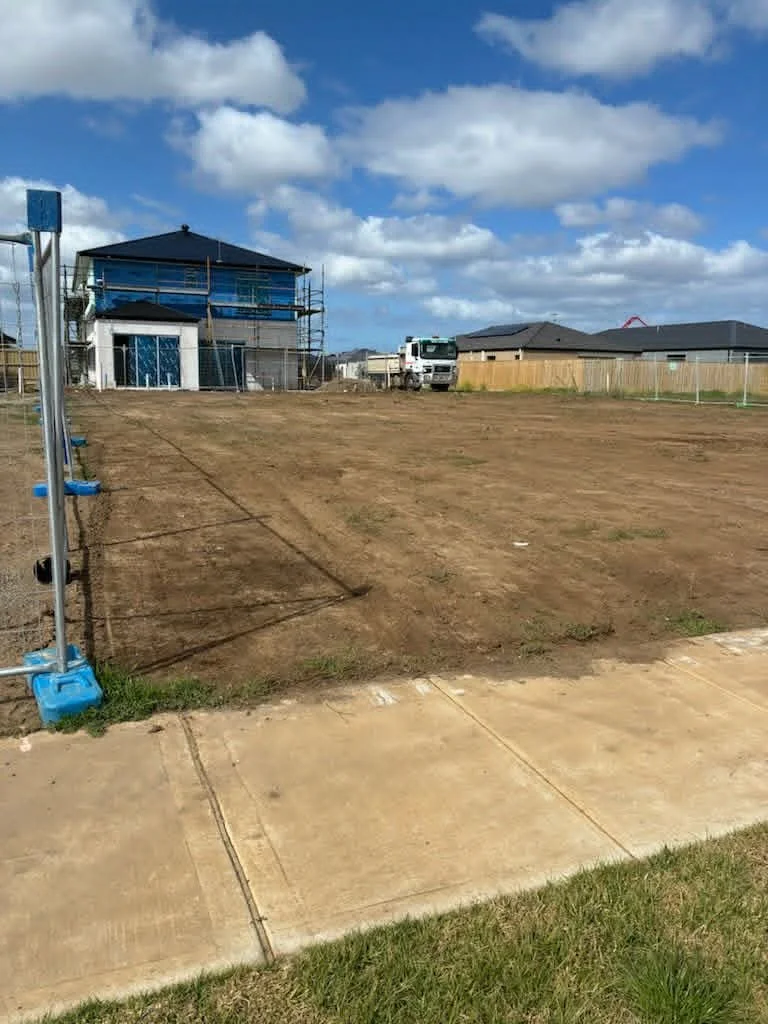 Empty lot with dirt, a sidewalk in the foreground, and a house under construction in the background. The house has blue siding and scaffolding around it, with some construction vehicles parked nearby. The sky is partly cloudy.