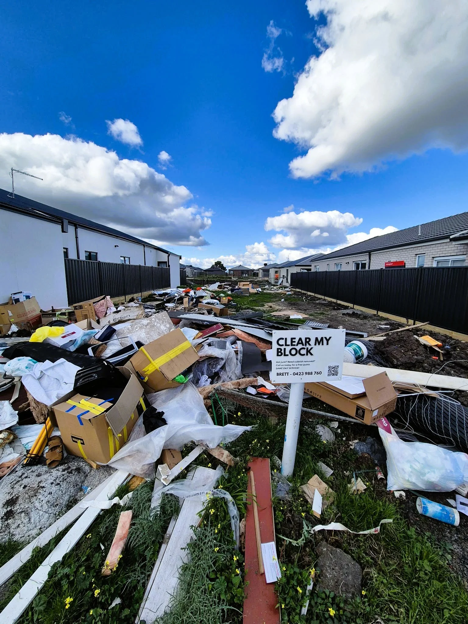 A residential street with scattered garbage and debris, including cardboard boxes, plastic bags, and construction materials, in front of newly built houses with black fences during daytime with a cloudy blue sky.