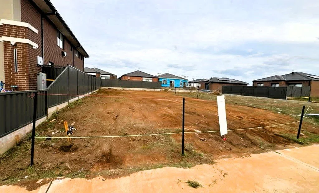 Vacant lot with cleared soil, surrounded by black fences, in a residential neighborhood with houses and a paved sidewalk.