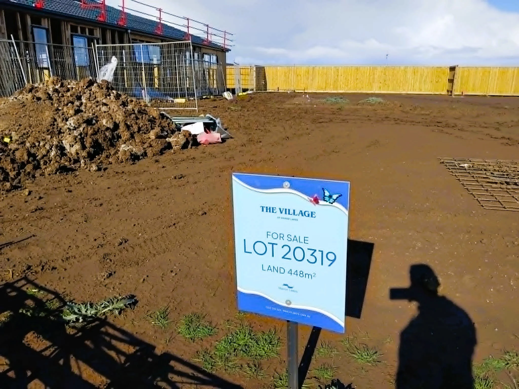 Empty plot of land enclosed with a yellow wooden fence, with a construction sign indicating lot number 20319 and land size 448 square meters in front. There is a pile of dirt on the left, some construction materials and equipment nearby.