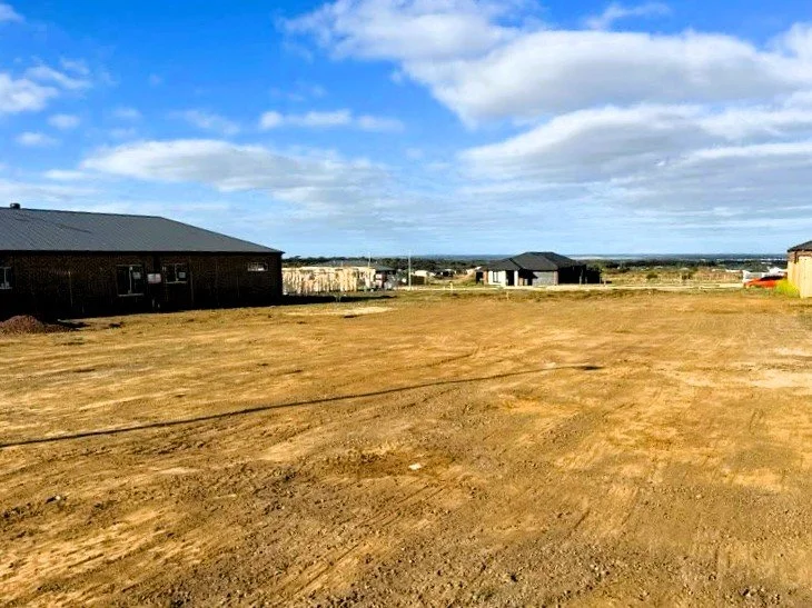 Empty dirt lot with scattered buildings in the background under a partly cloudy sky.