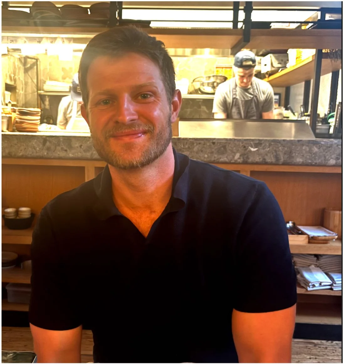 A man with short brown hair and a beard smiling at a restaurant or cafe with a kitchen visible in the background.
