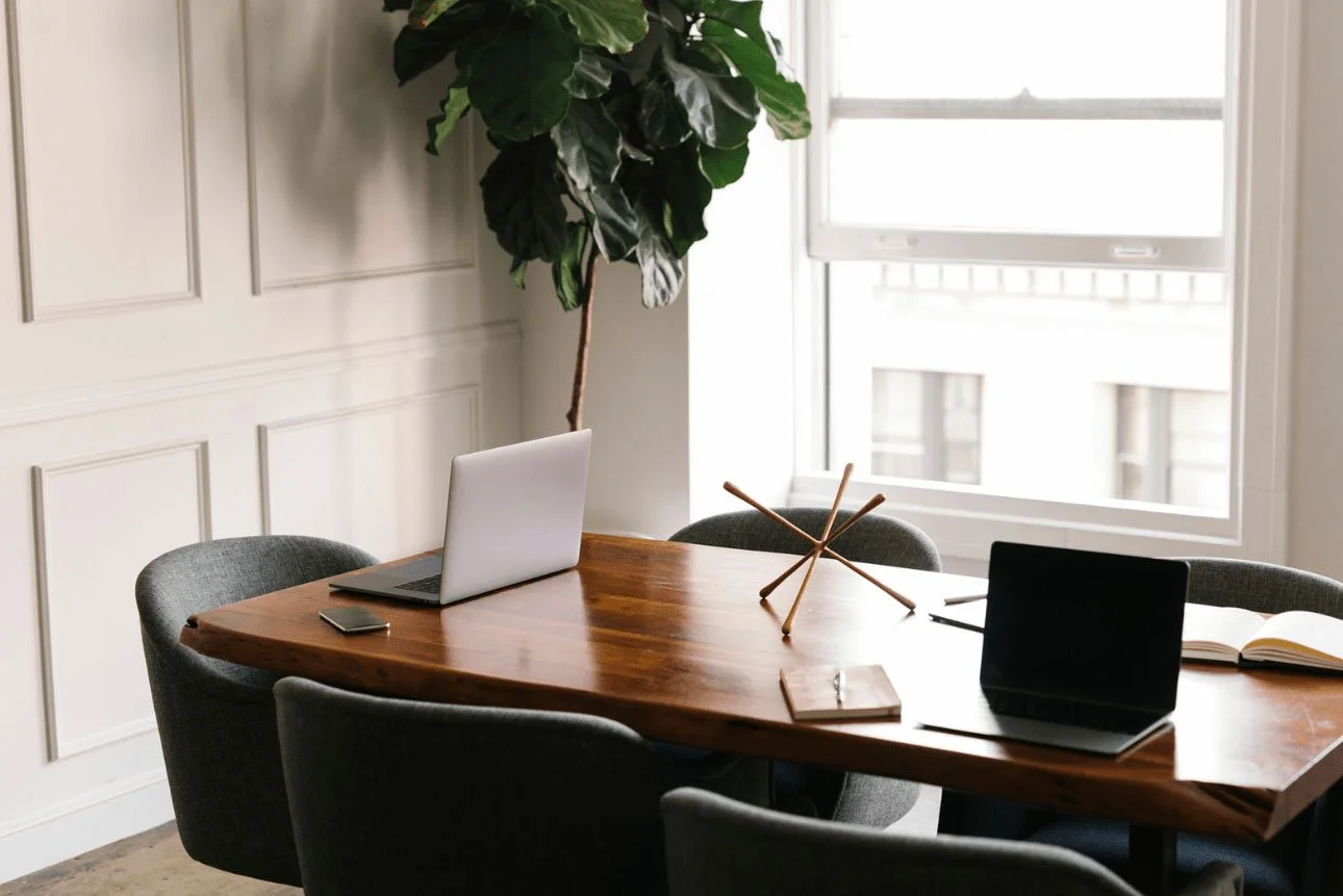 A modern office conference room with a wooden table, three gray chairs, a laptop, a tablet, a notebook, a large plant, and a window with blinds.