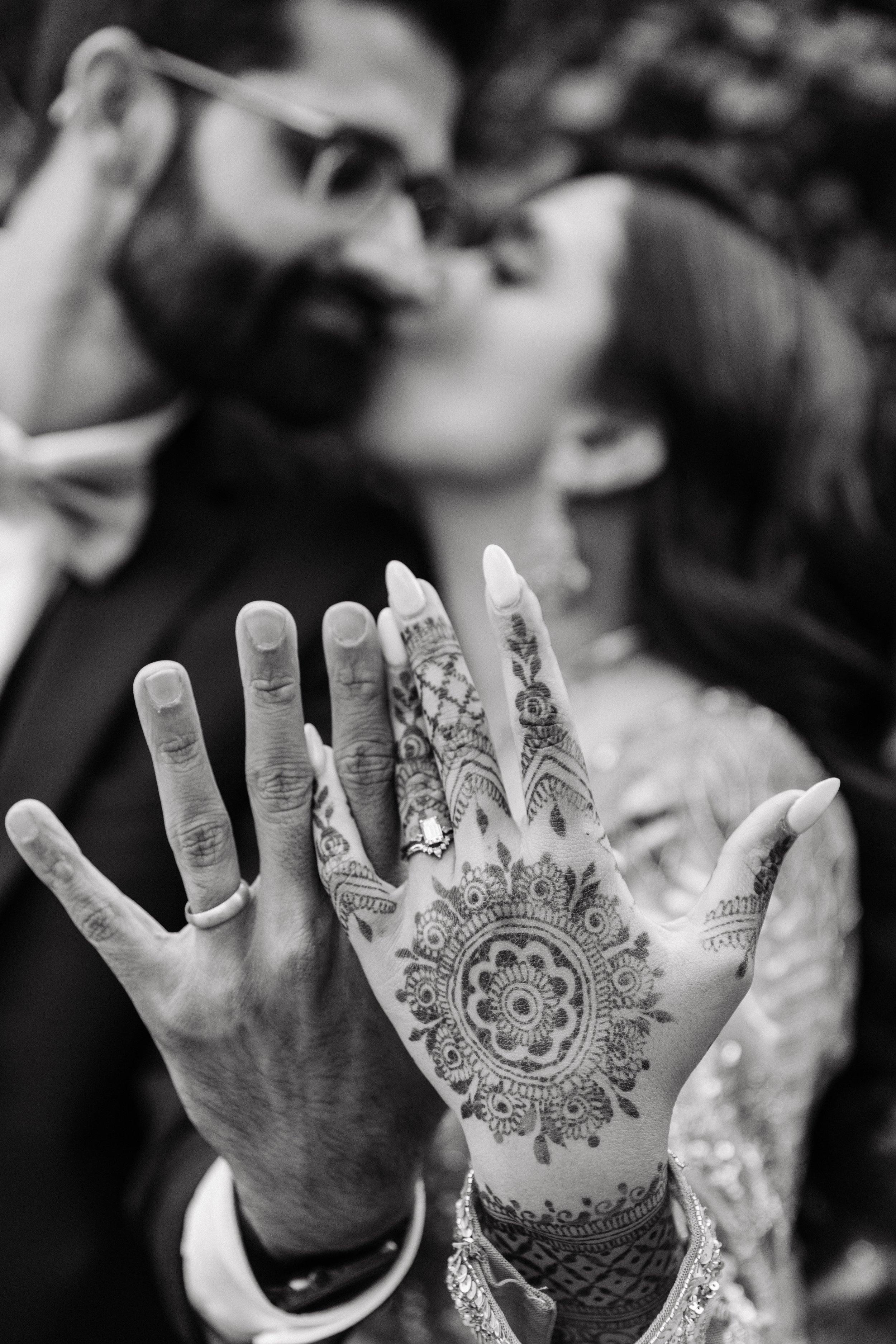 Close-up of a couple showing their hands with henna designs and wedding rings, blurry background of them kissing.
