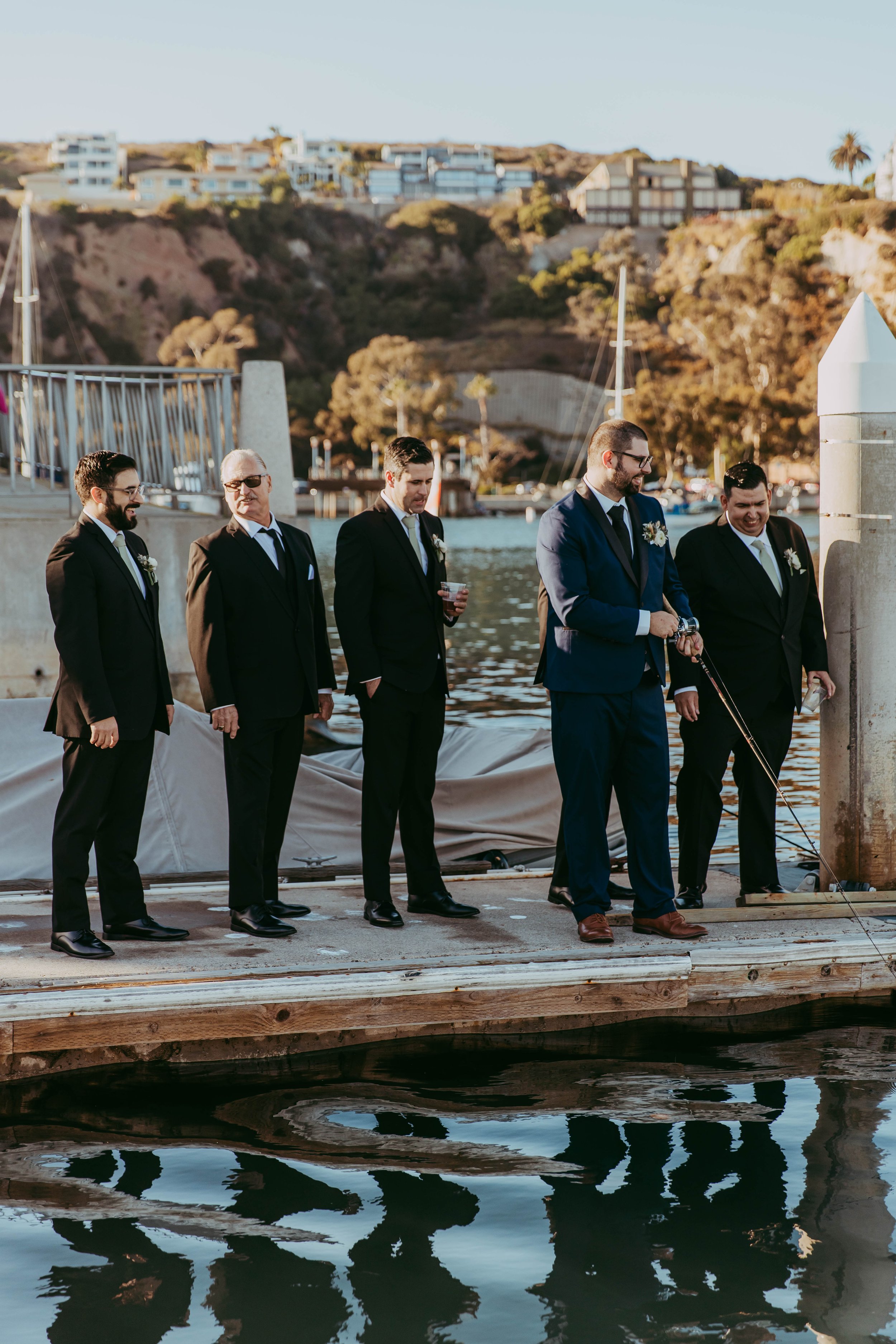 Group of men in formal suits standing on a dock by the water, with a scenic hillside and houses in the background.