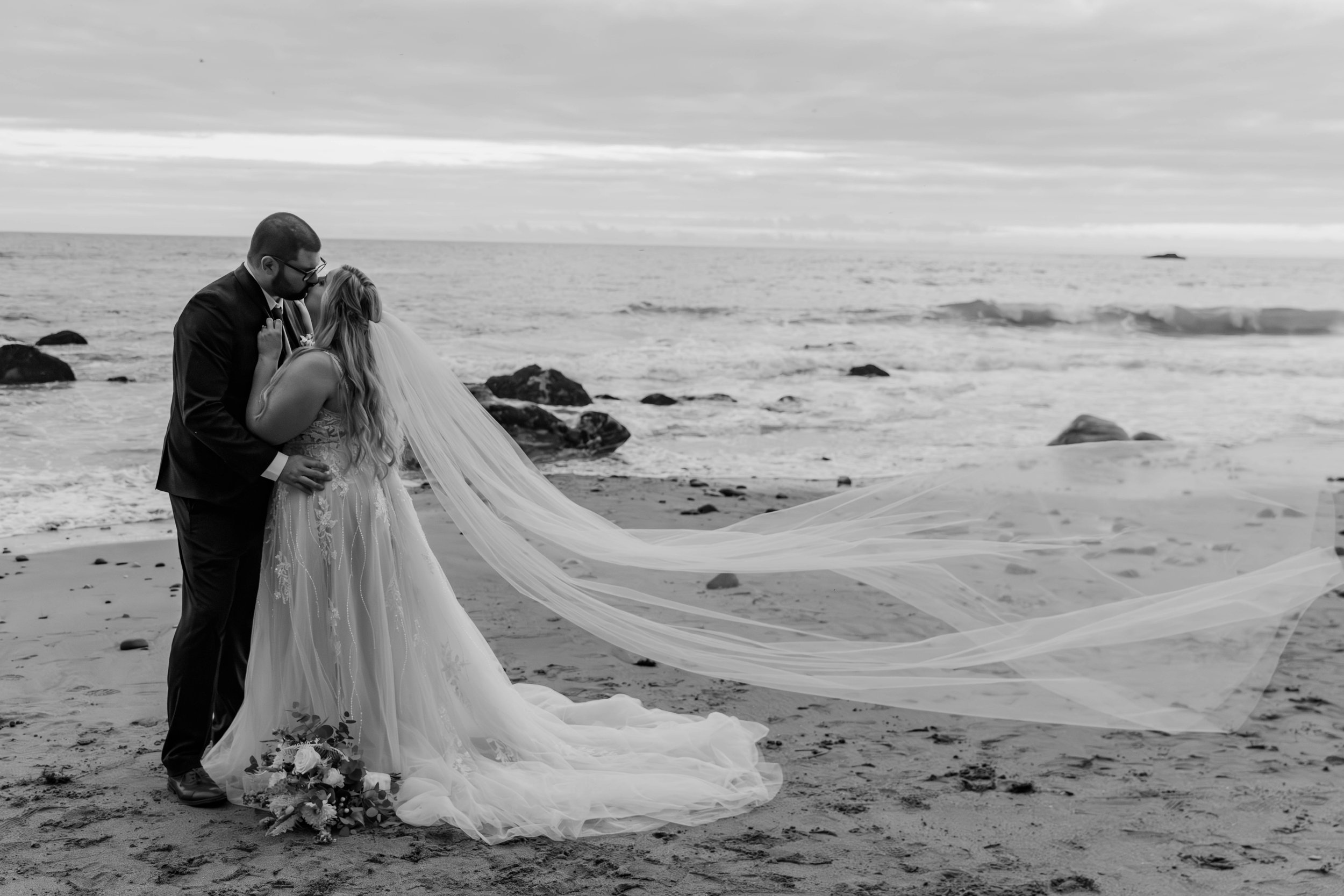 Black and white photo of a bride and groom kissing on a beach with a long flowing veil and bouquet on the sand.