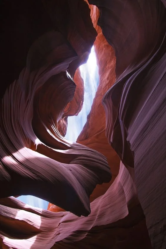 Inside a narrow slot canyon with smooth, layered red and purple rock walls and a beam of sunlight shining down from above.