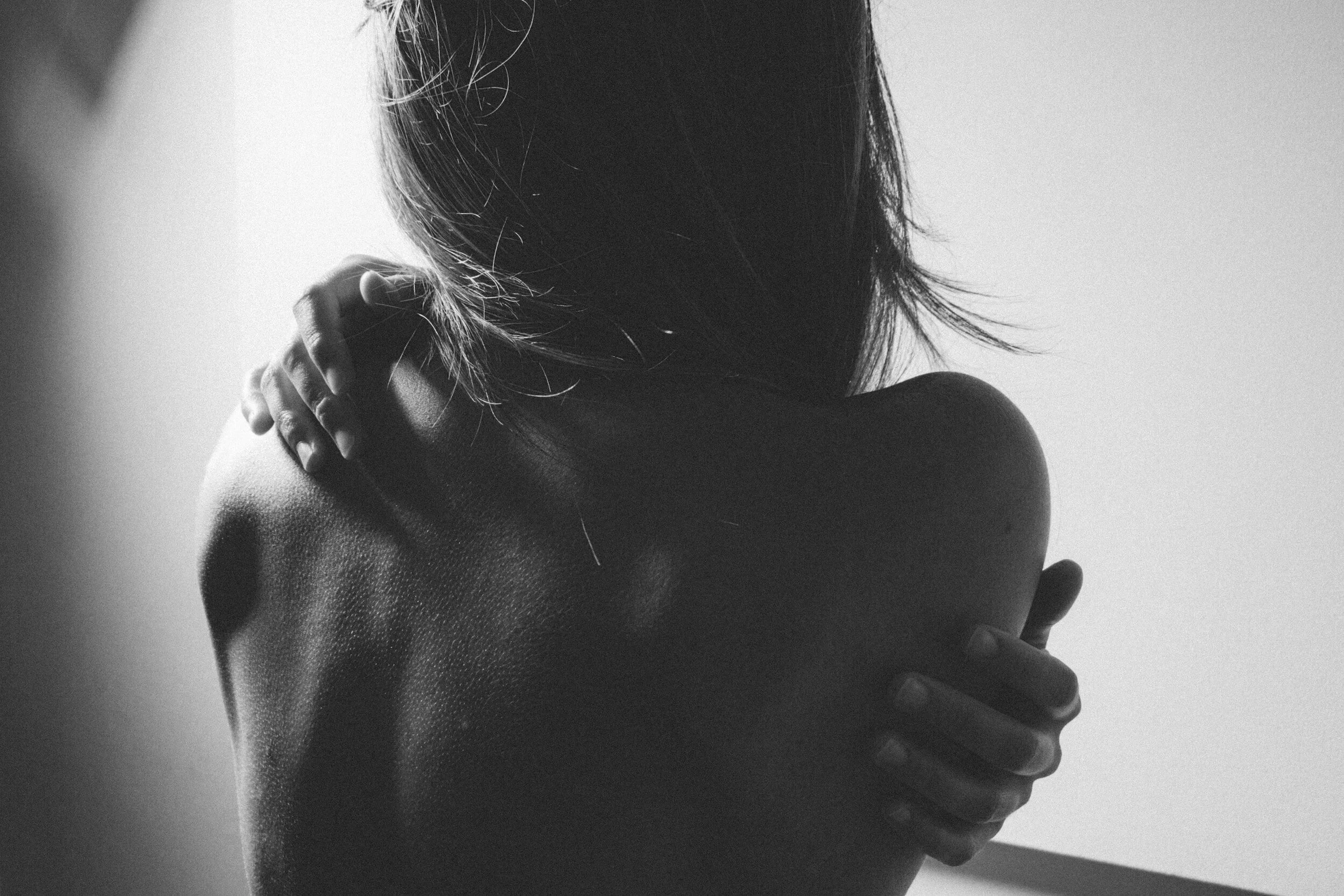 Black and white photograph of a woman hugging herself, with her arms crossed over her chest and her head turned away, focused on her bare shoulders and back.