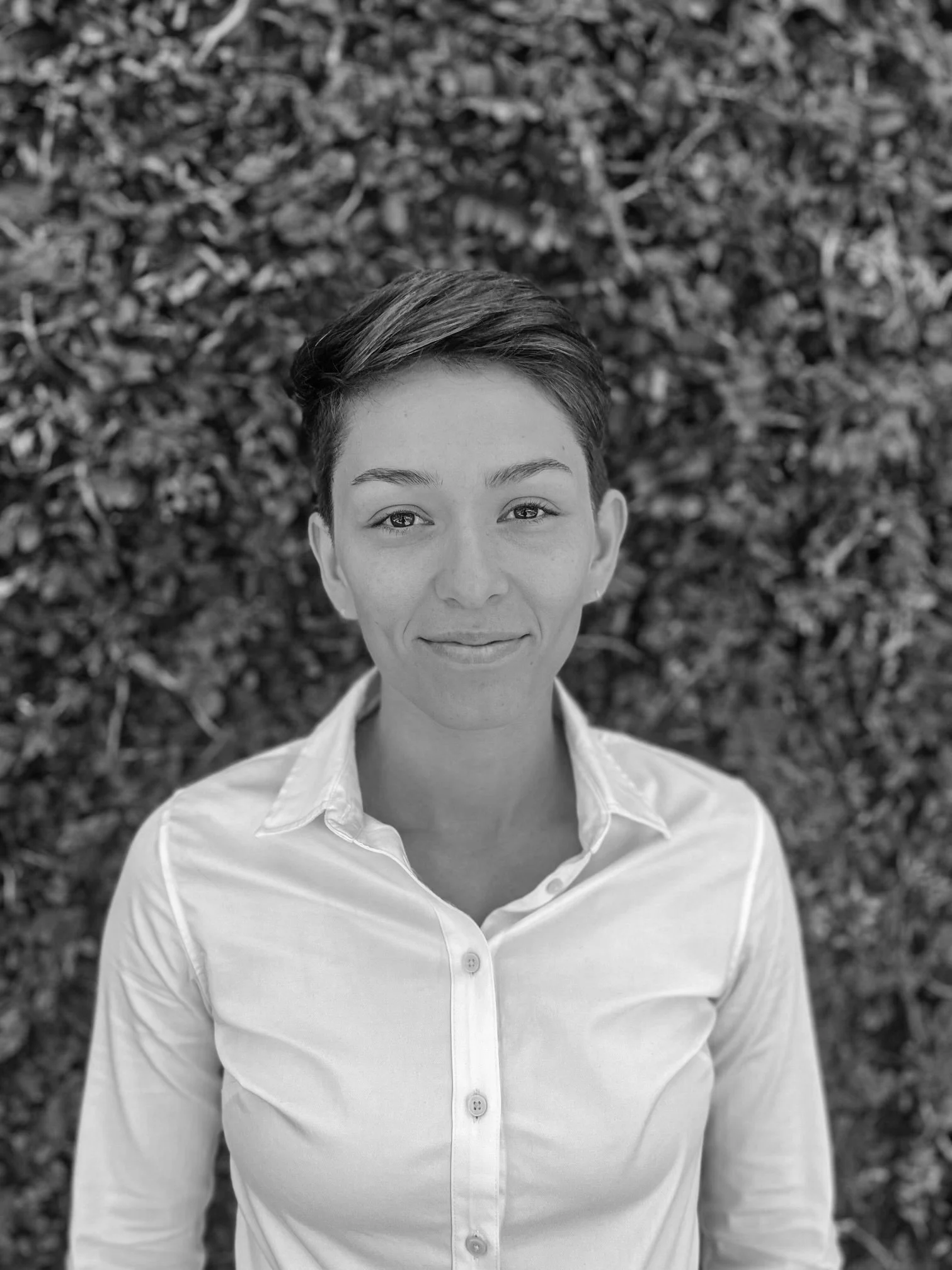Black and white portrait of a woman with short hair, wearing a button-up shirt, standing outdoors in front of a leafy background.