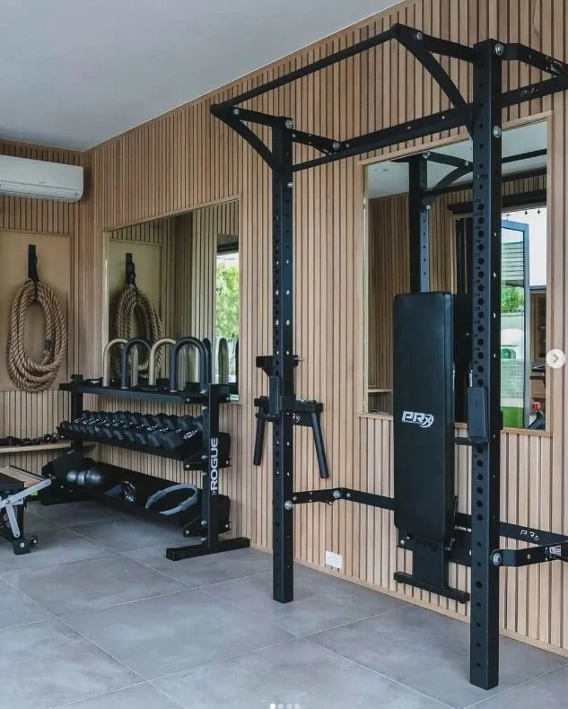 Home gym with a black power rack, weight bench, dumbbells, kettlebells, medicine balls, and wall-mounted ropes, all set against wood-paneled walls.