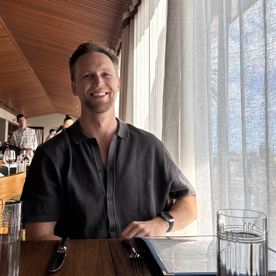 A man sitting at a restaurant table near a large window with curtains, smiling, with a knife and fork on the table, and other people dining in the background.