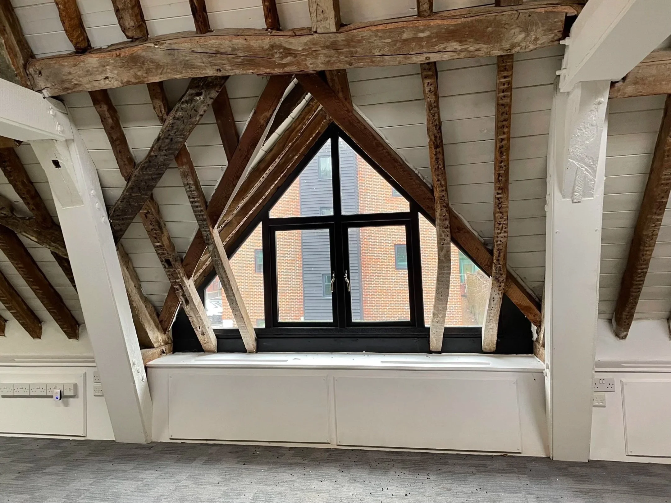 Interior view of a room with exposed wooden roof beams and a triangular window showing a brick building outside.