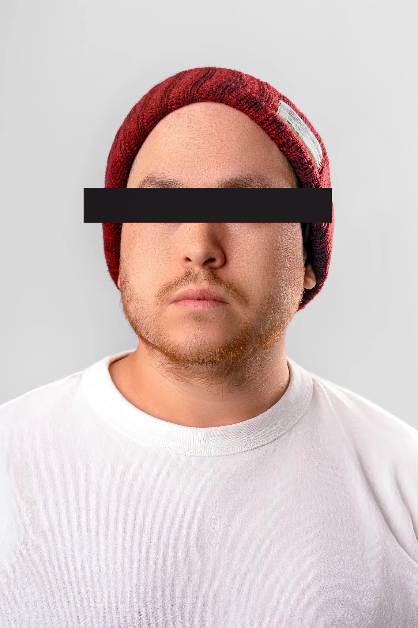 A young man with a red knit beanie, wearing a white t-shirt, has a black bar obscuring his eyes in this portrait photo.