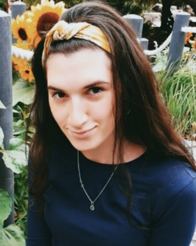 A woman with long brown hair wearing a yellow headband and a navy blue top, standing in front of sunflowers and greenery.