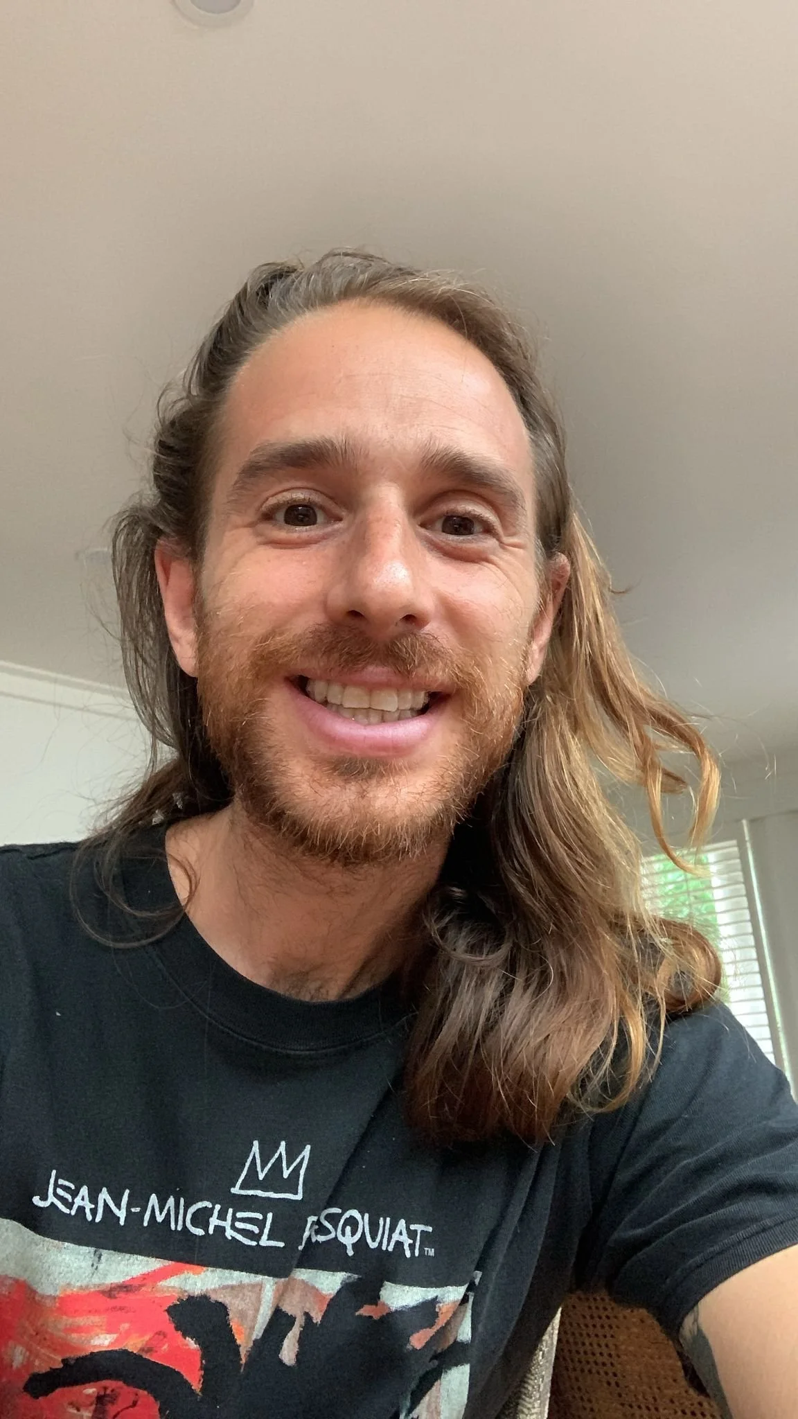 A smiling man with long, wavy hair and a beard wearing a black Jean-Michel Basquiat T-shirt indoors.
