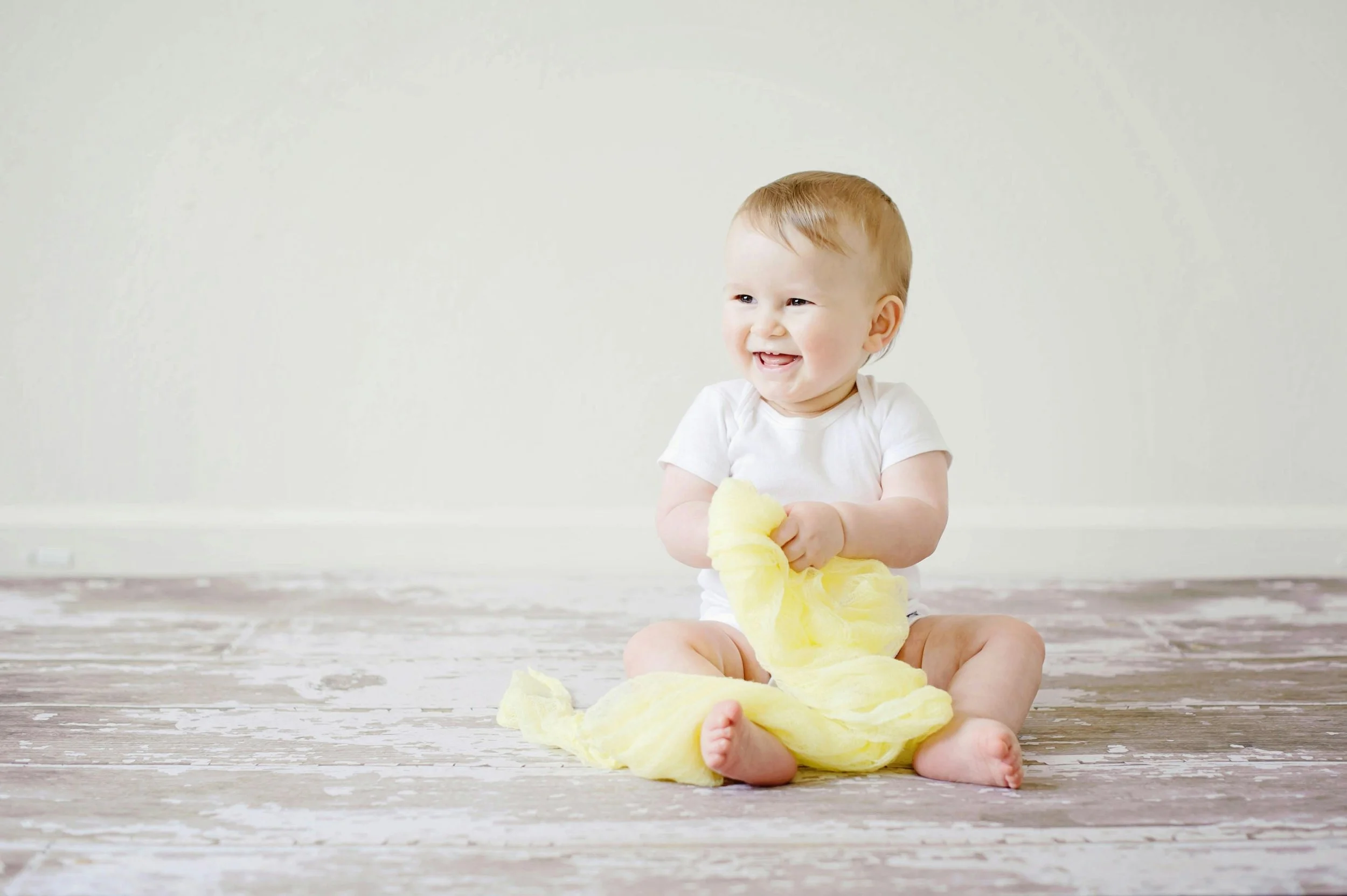 Lächelndes Baby sitzt auf einem Holzboden und spielt mit einem gelben Tuch.