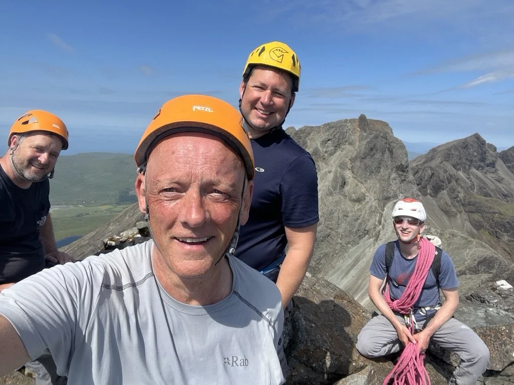 Four climbers wearing helmets and harnesses on a mountain summit with rocky peaks and a valley in the background.