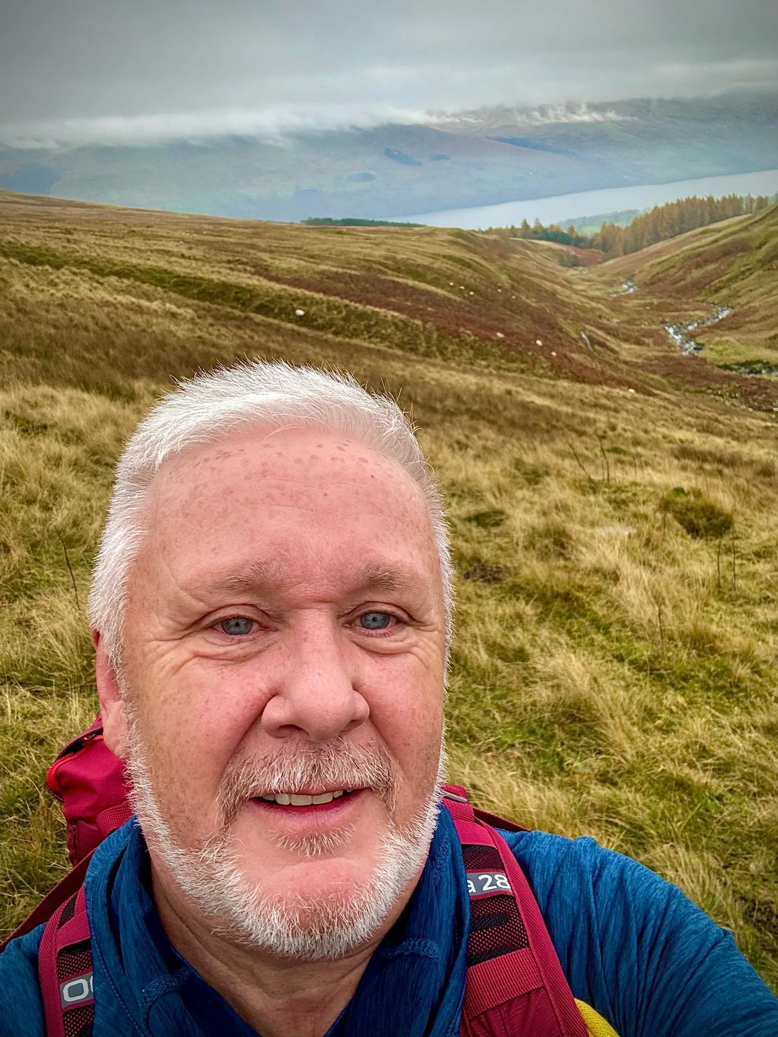 A man with white hair and beard taking a selfie in a grassy, hilly landscape with a lake and cloudy sky in the background.
