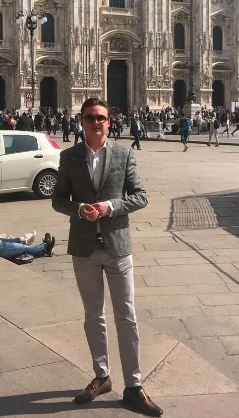 A man in a plaid blazer and sunglasses standing on a city street with a historic building in the background, surrounded by crowds and cars.