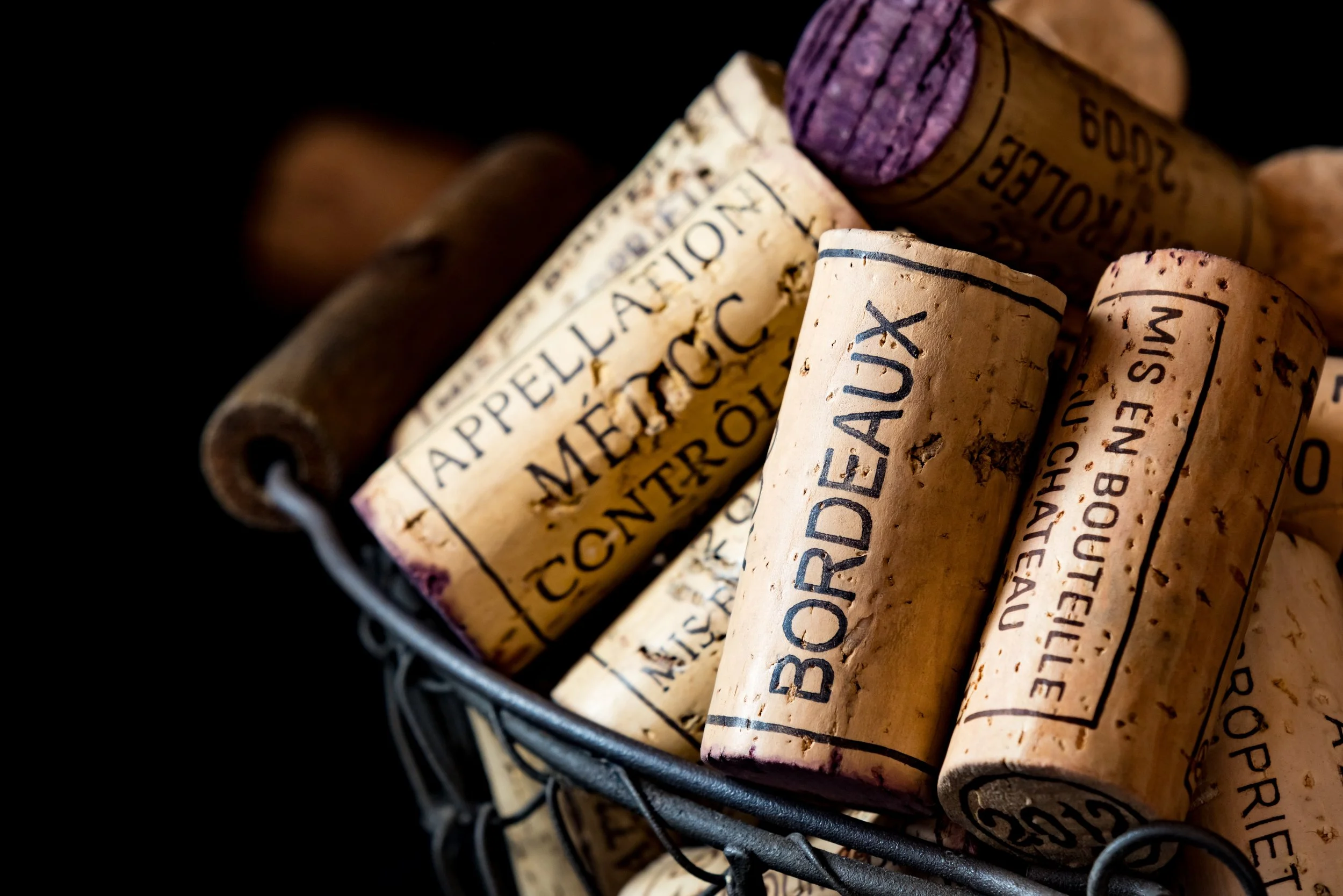A wire basket filled with used wine corks with handwritten and printed labels, including names like Bordeaux and Saint-Emilion.