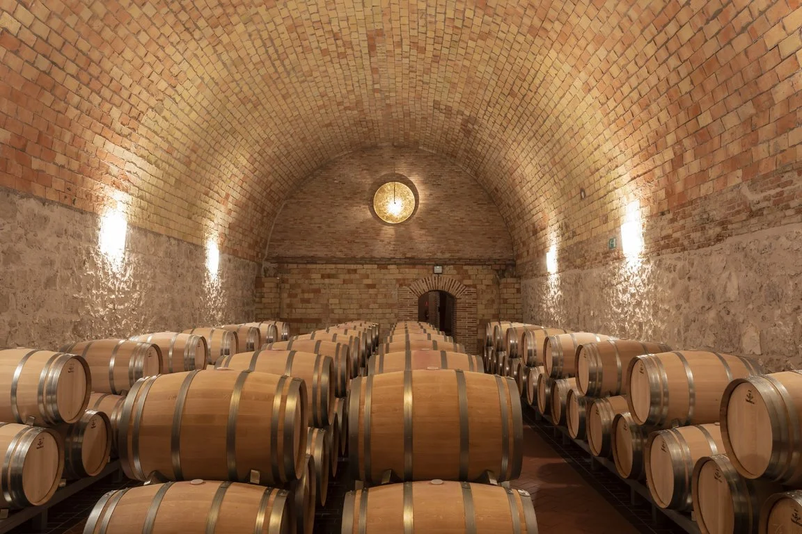 Rows of wine barrels stored in a wine cellar with exposed brick walls and a vaulted ceiling.