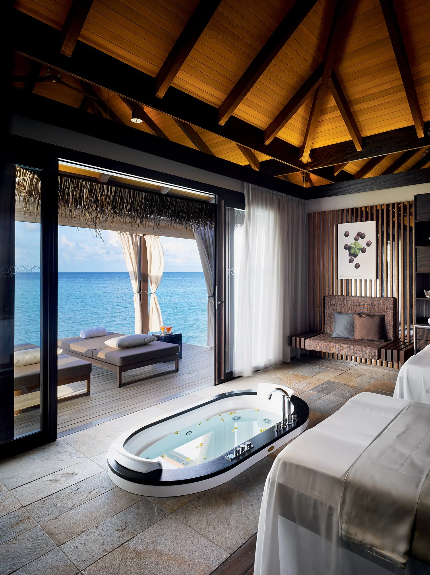 Luxurious hotel room with a view of the ocean, featuring a hot tub, a bed, sitting area with sofa, and balcony with lounge chairs.