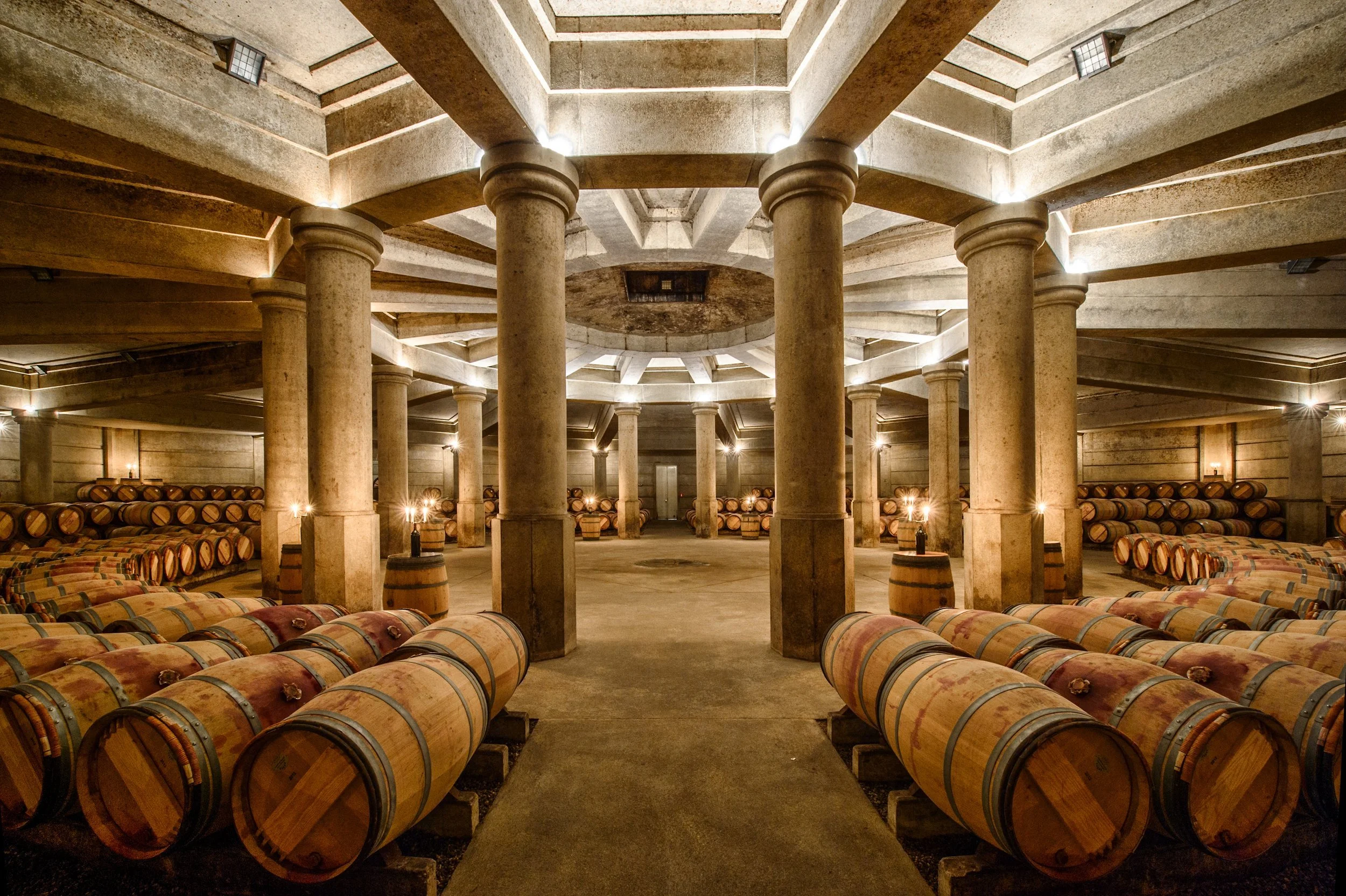 A large wine cellar with numerous stacked wine barrels on the ground and shelves, with stone columns and soft lighting.