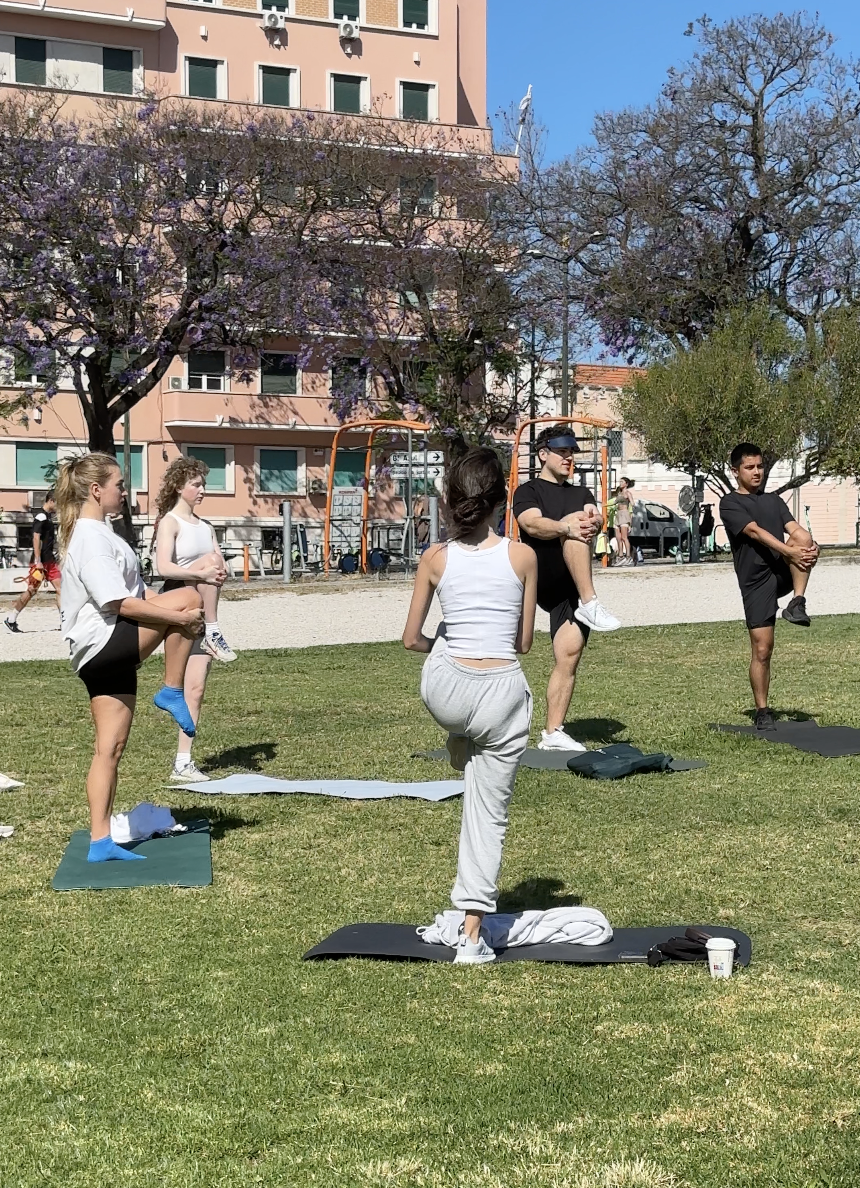 Group of people doing outdoor exercise on a grassy field, with yoga mats, in front of a building and trees.