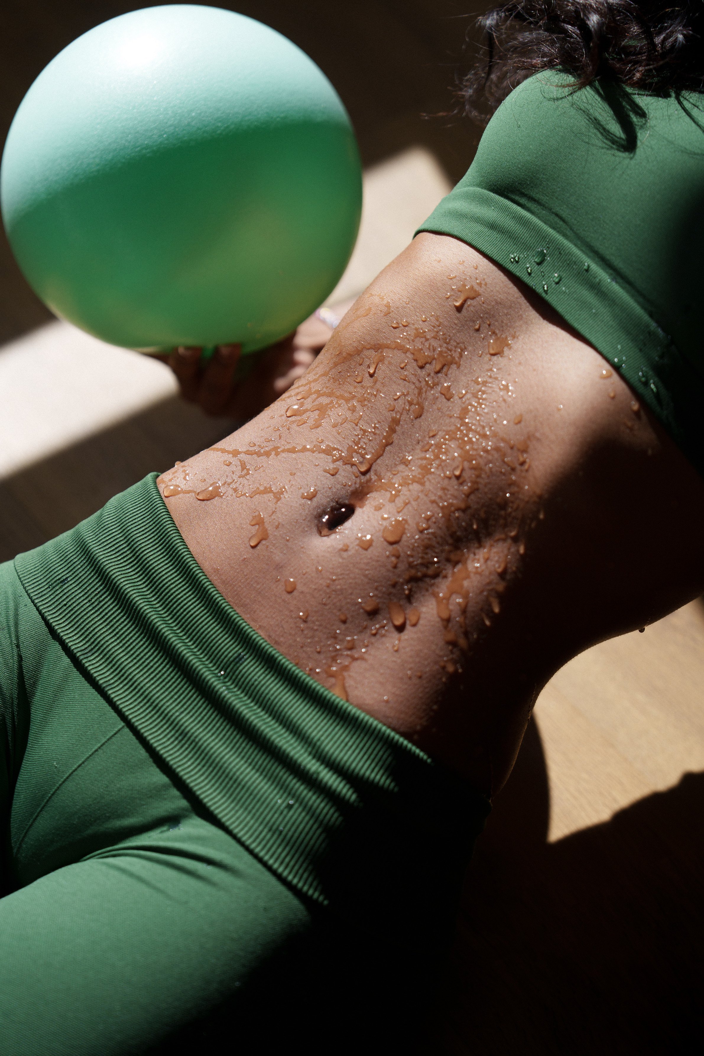 Close-up of a person's midsection wearing green workout clothing, holding a green exercise ball, with sweat on their skin, indicating fitness activity.