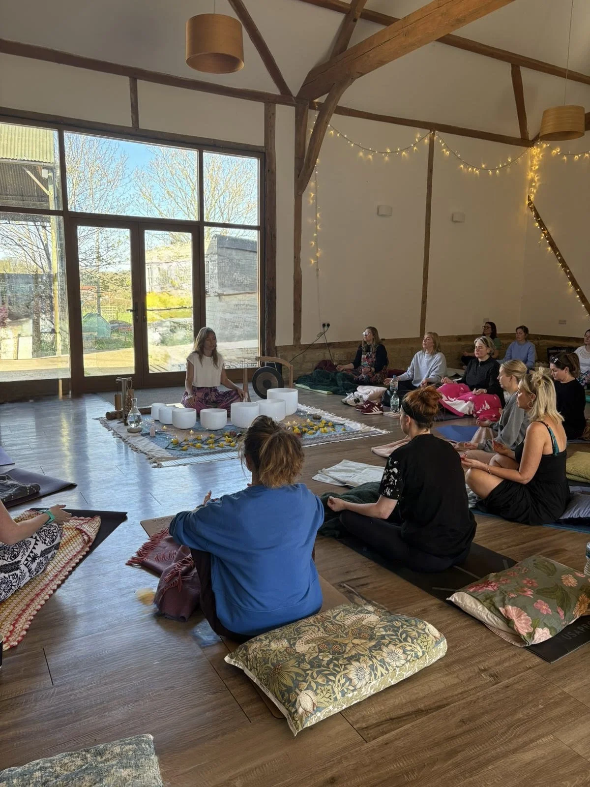 A group of people participating in a meditation or yoga session inside a spacious, well-lit room with large windows and wooden beams. They are sitting on cushions on the floor, facing a woman seated in front who is leading the session. The room is decorated with fairy lights.
