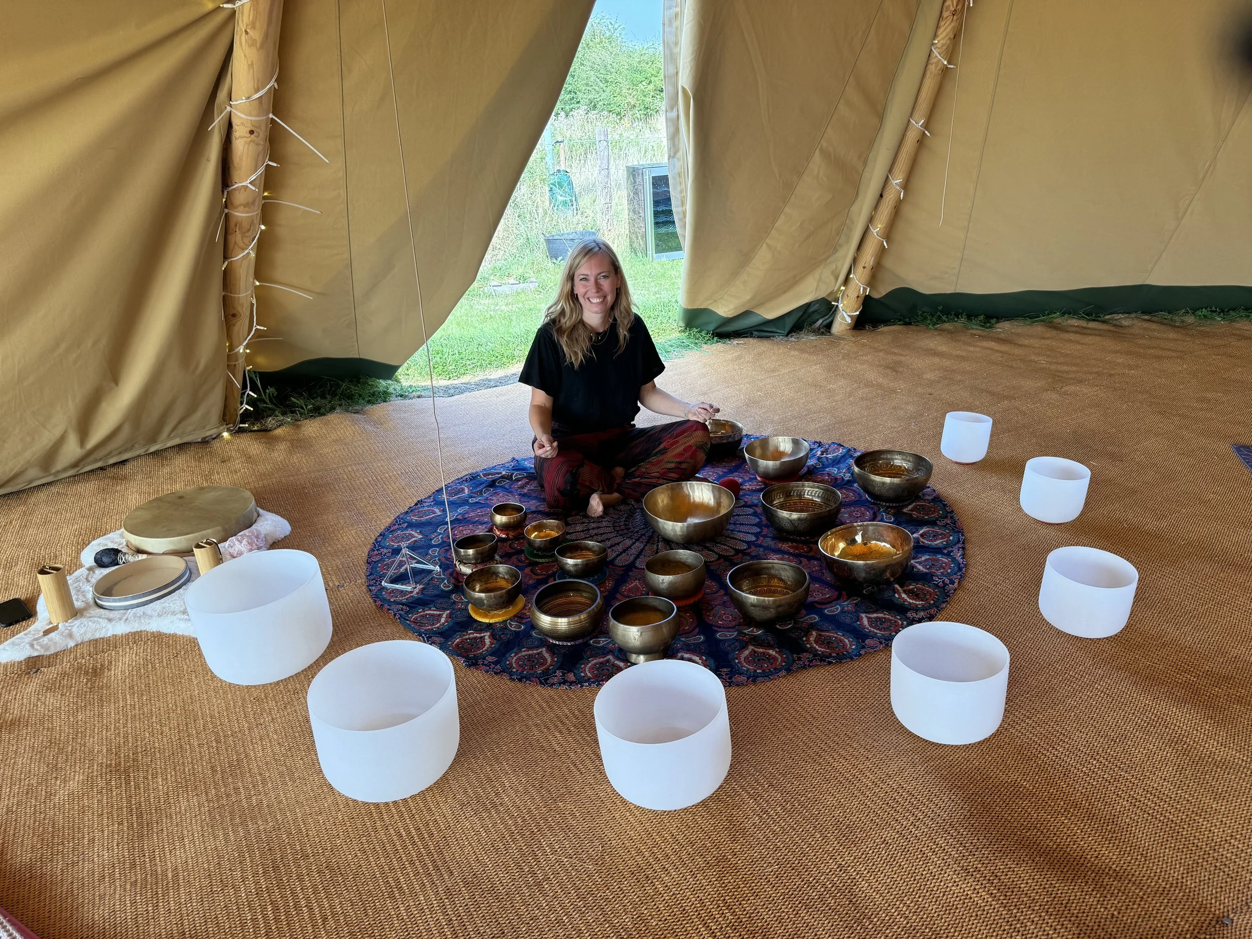 A woman sitting cross-legged inside a large beige tent, surrounded by a colorful circular rug and several metallic singing bowls of various sizes, with some white crystal singing bowls and a round cushion nearby, outdoor greenery visible through the tent opening.
