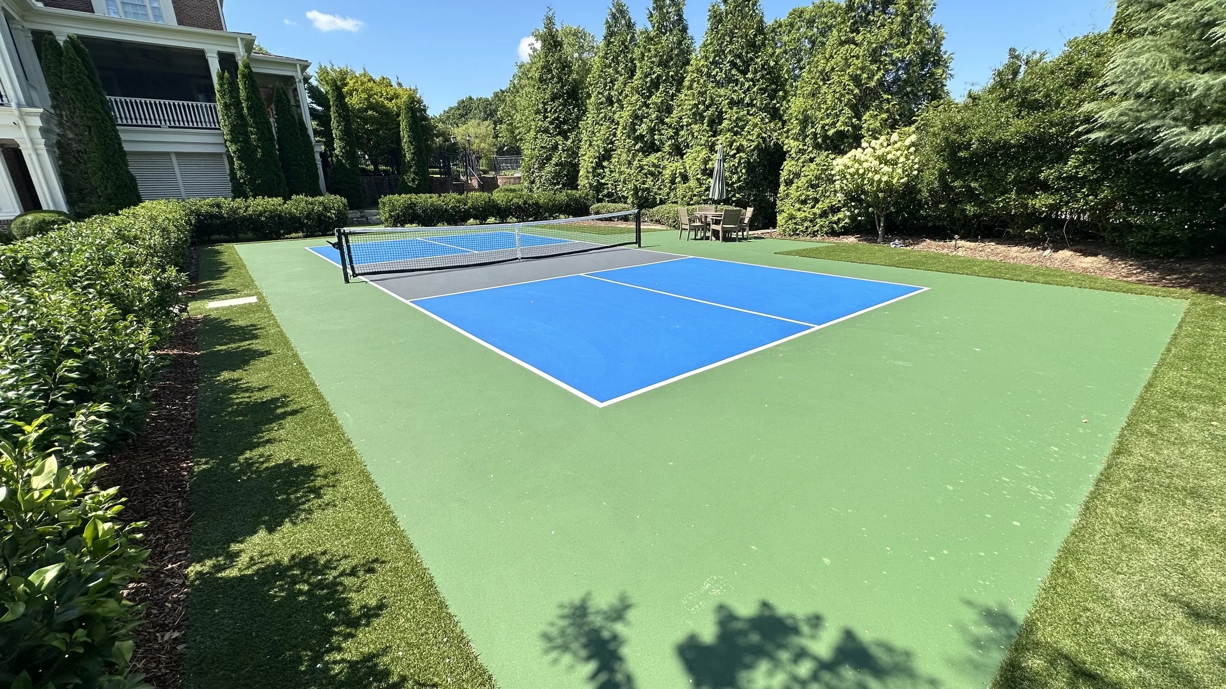 An outdoor backyard with a blue and green tennis or pickleball court, surrounded by lush green trees and shrubs, with a patio area and umbrella in the background.