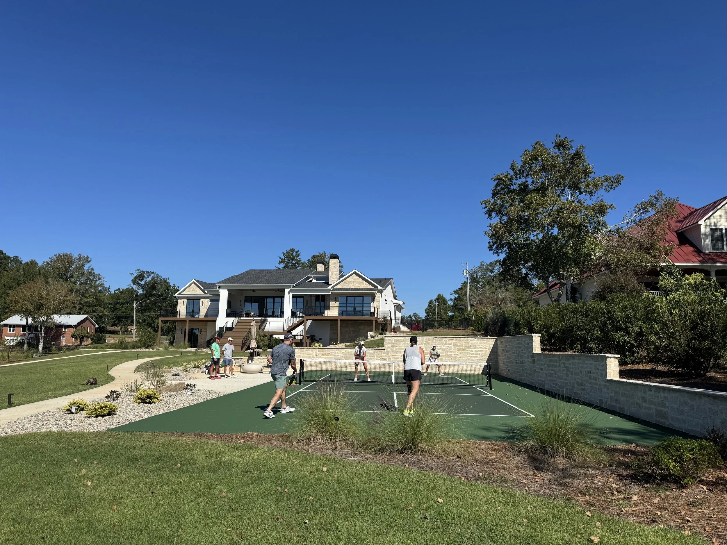 Group of people playing tennis on an outdoor court in a residential area with large houses and trees in the background, under a clear blue sky.
