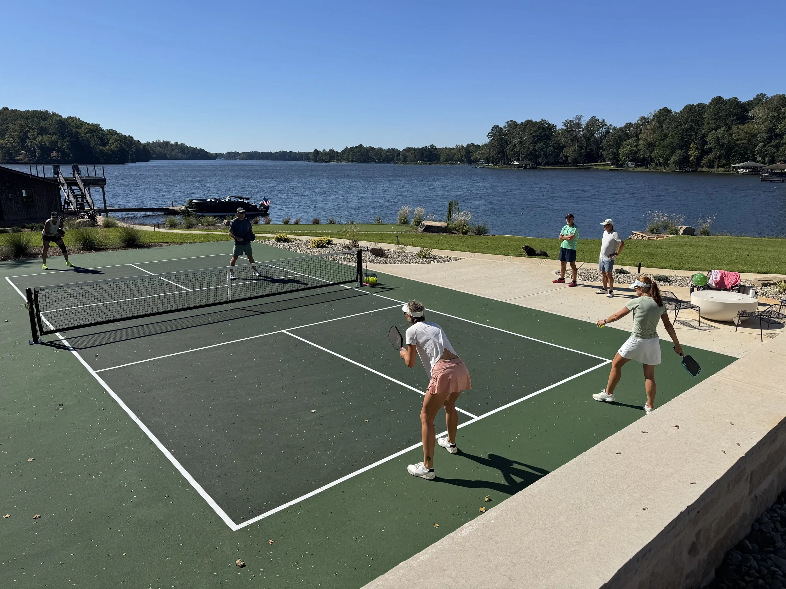 People playing pickleball on an outdoor court beside a lake with trees and a boat dock in the background.