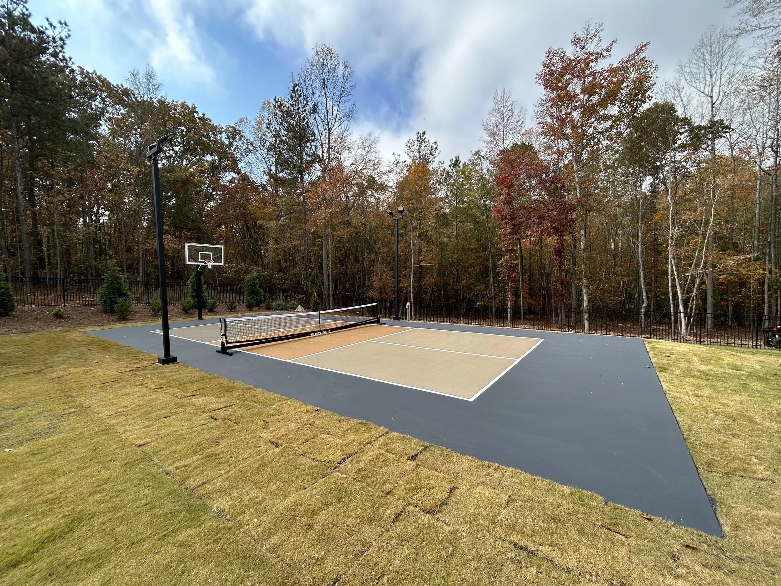 A newly built outdoor pickleball court with a tan playing surface, surrounded by a darker blue border, fenced in, with two basketball hoops and a net, situated in a wooded area during fall with colorful trees in the background.