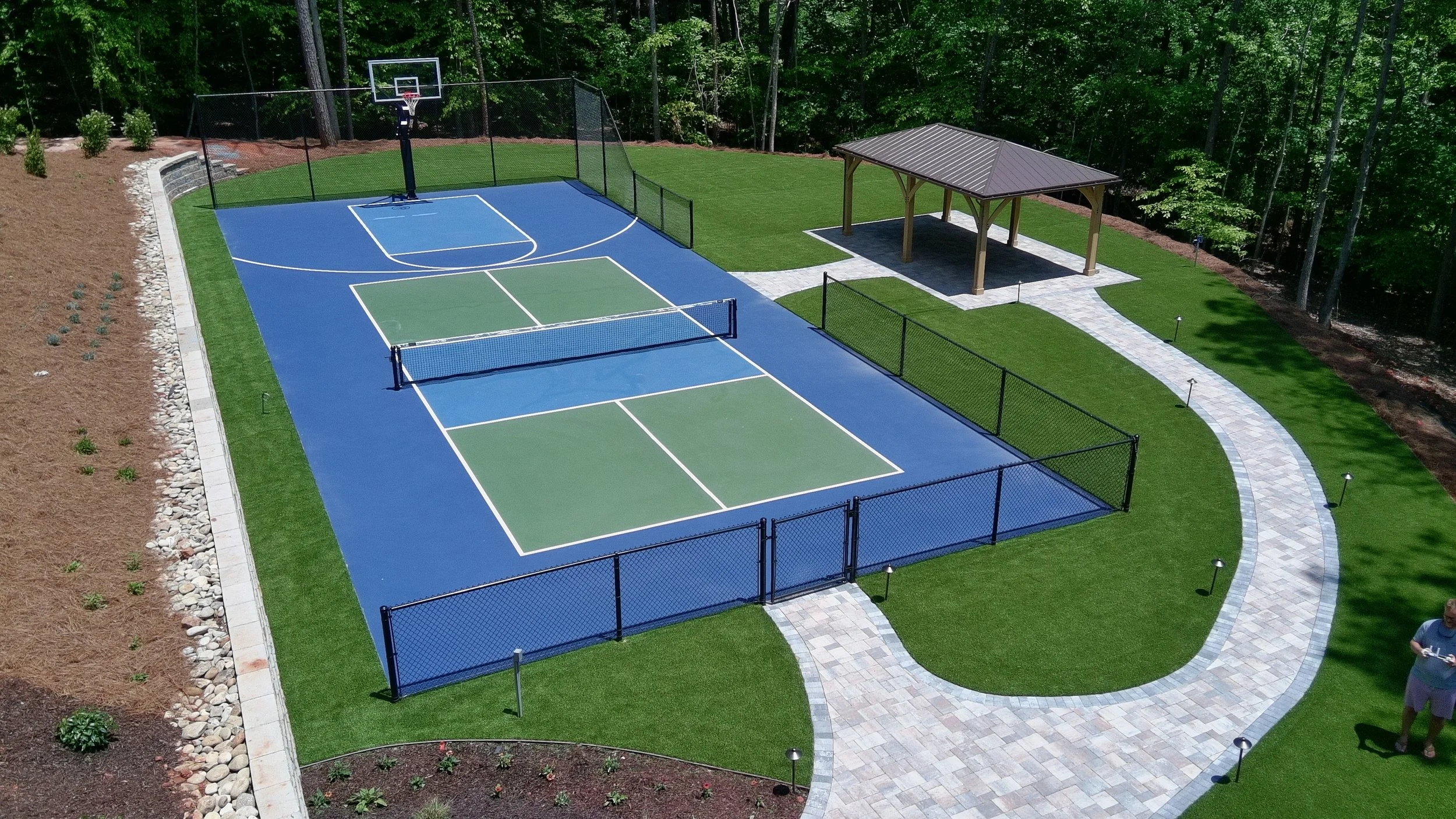 An outdoor sports court with a basketball hoop, a volleyball net, and a small covered pavilion surrounded by trees and landscaped landscaping.