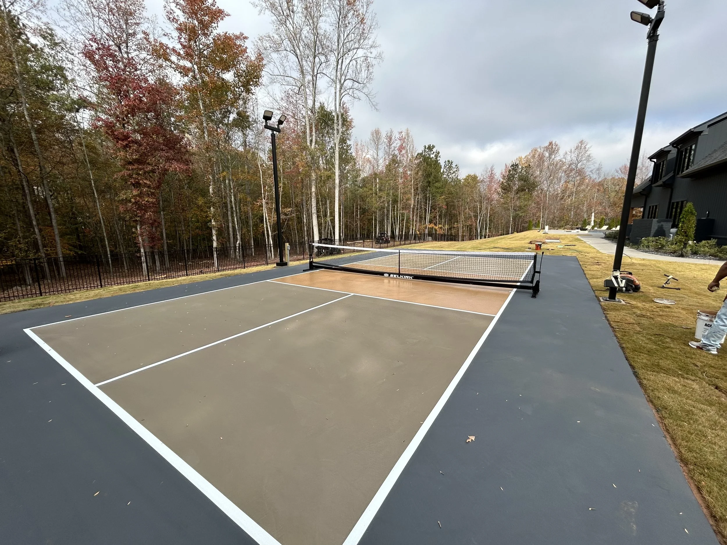 An outdoor pickleball court with a net and painted lines, surrounded by a black fence and tall trees in the background, with a cloudy sky overhead.