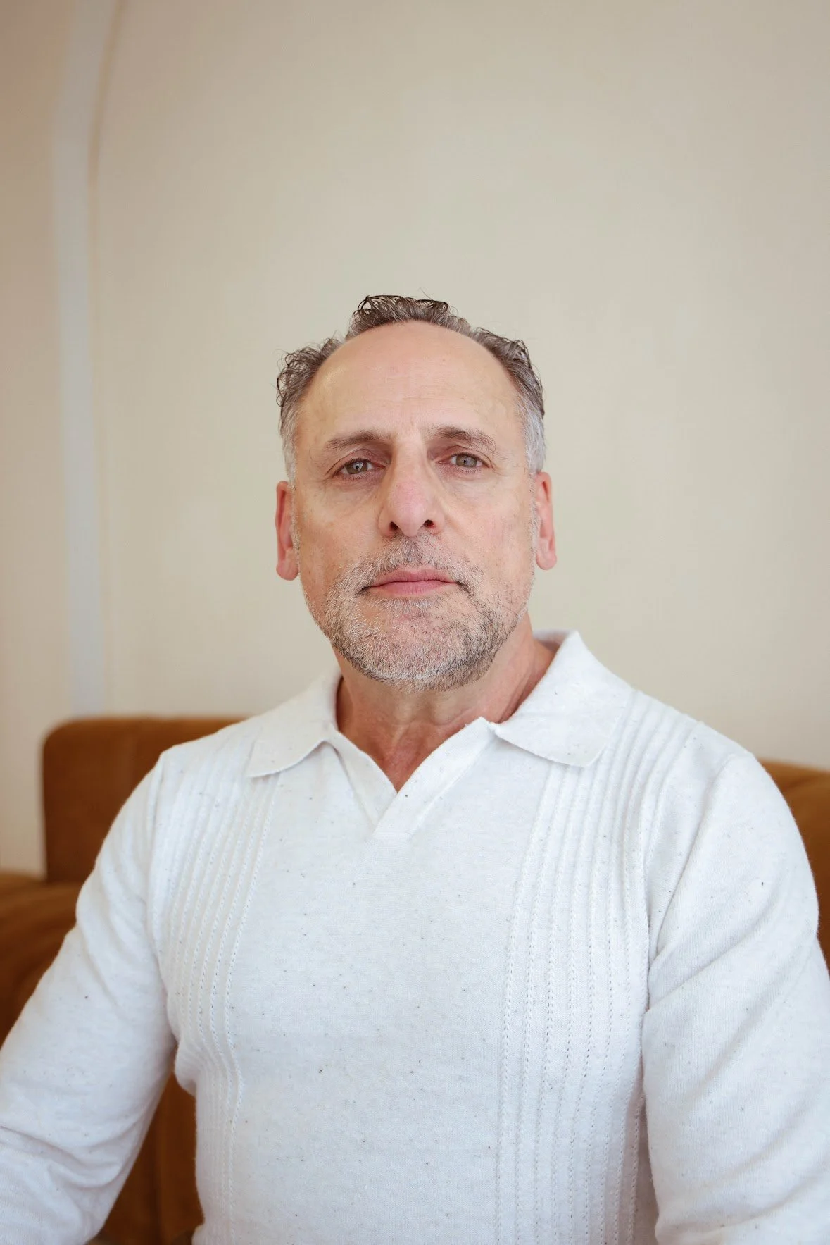 A middle-aged man with short, curly gray hair and a beard, wearing a white sweater, sitting on a brown couch against a plain, light-colored wall.