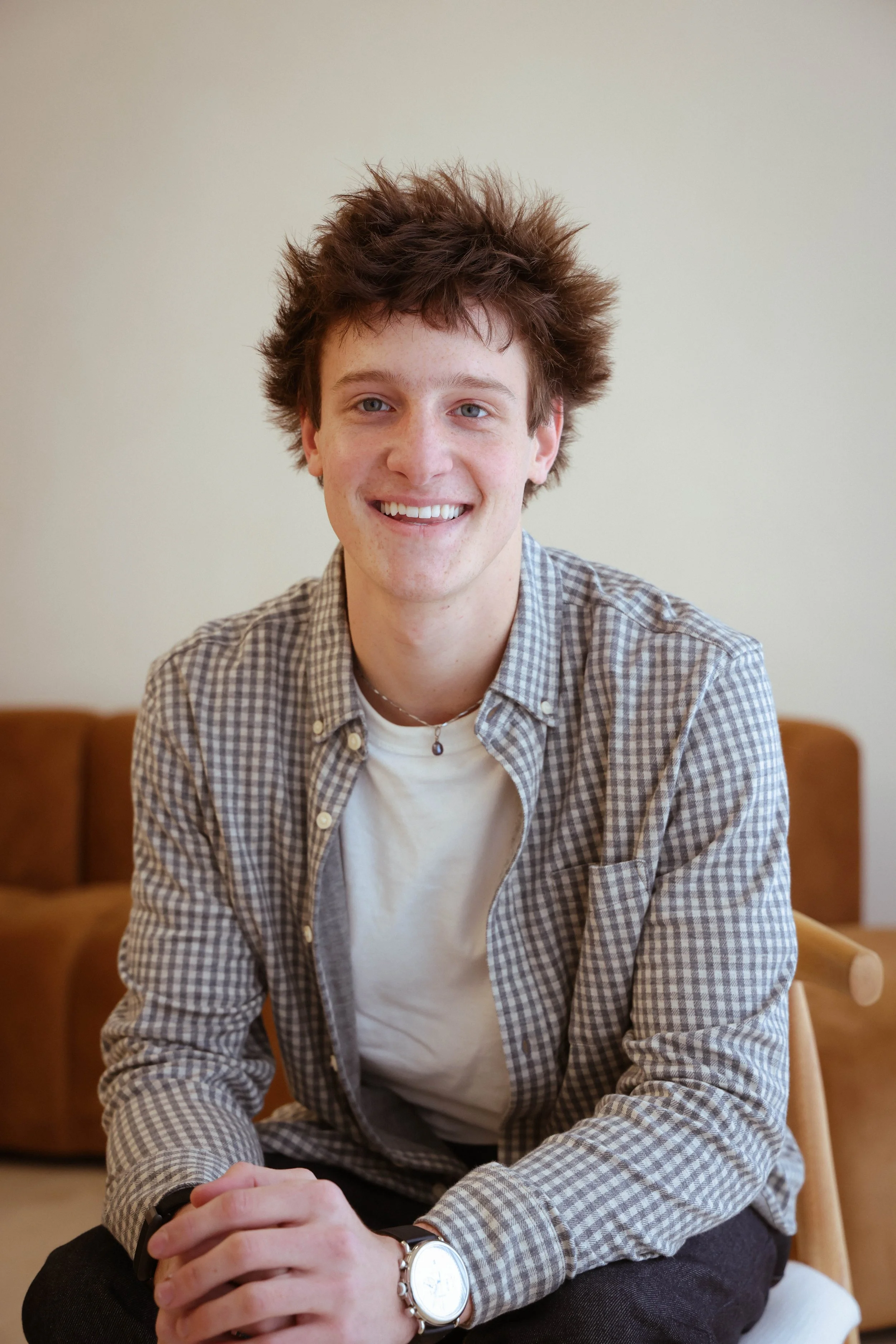 A young man with light skin, brown curly hair, wearing a checkered shirt over a white t-shirt, smiling while sitting with hands clasped in front of him in a room with a beige wall and sofa in the background.