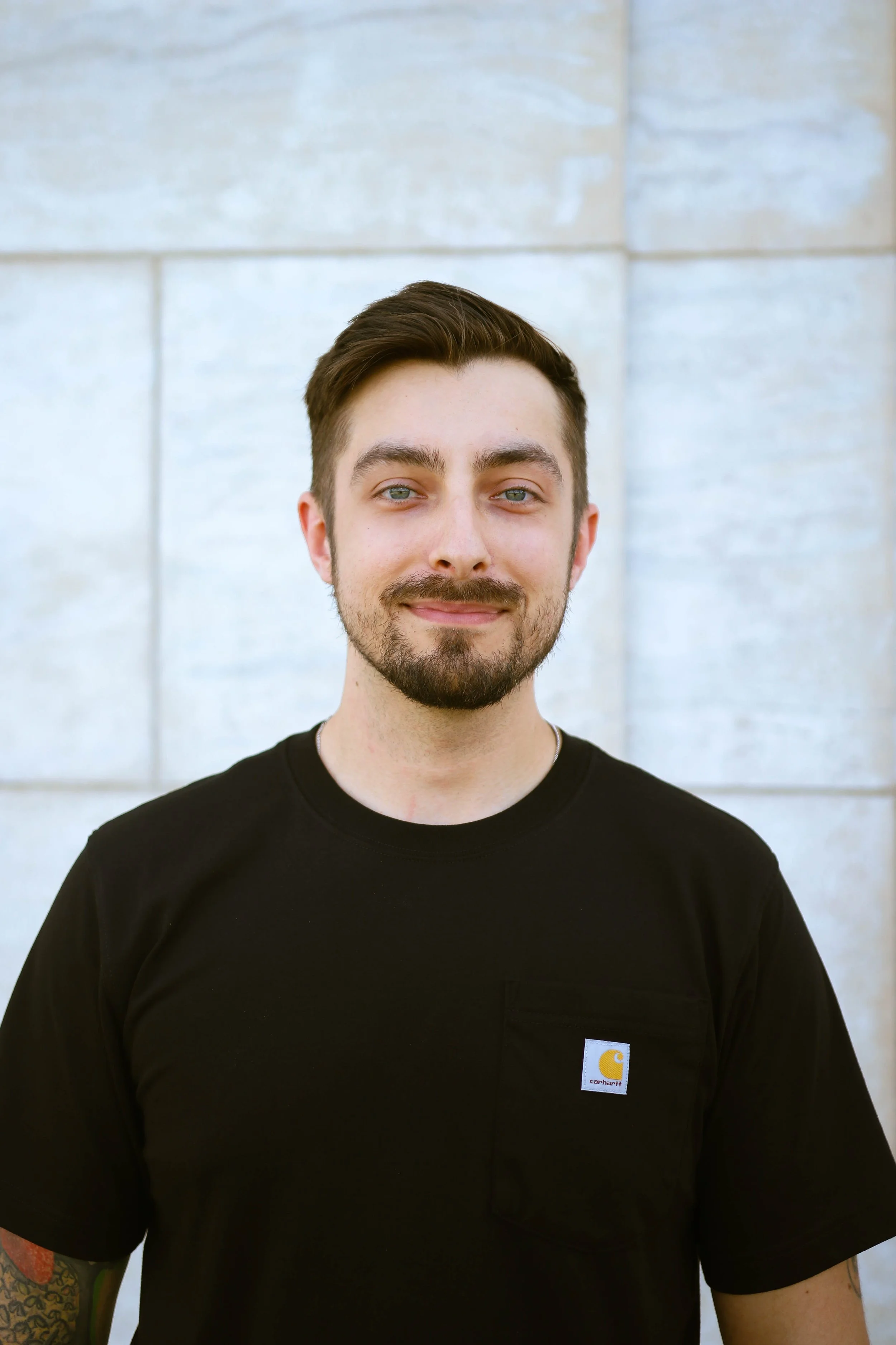 A young man with short brown hair, a beard, and blue eyes, wearing a black Carhartt t-shirt, standing outdoors in front of a light-colored stone wall.
