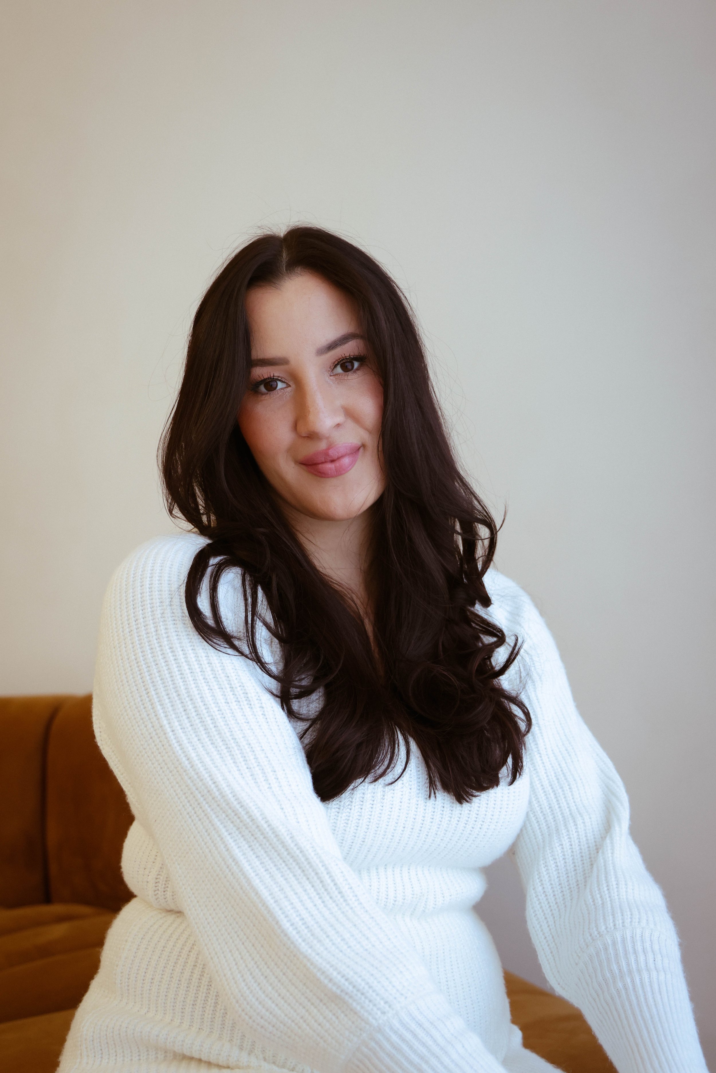 A woman with long dark hair, wearing a white knit sweater, sitting on a brown couch against a plain light-colored wall, smiling slightly at the camera.