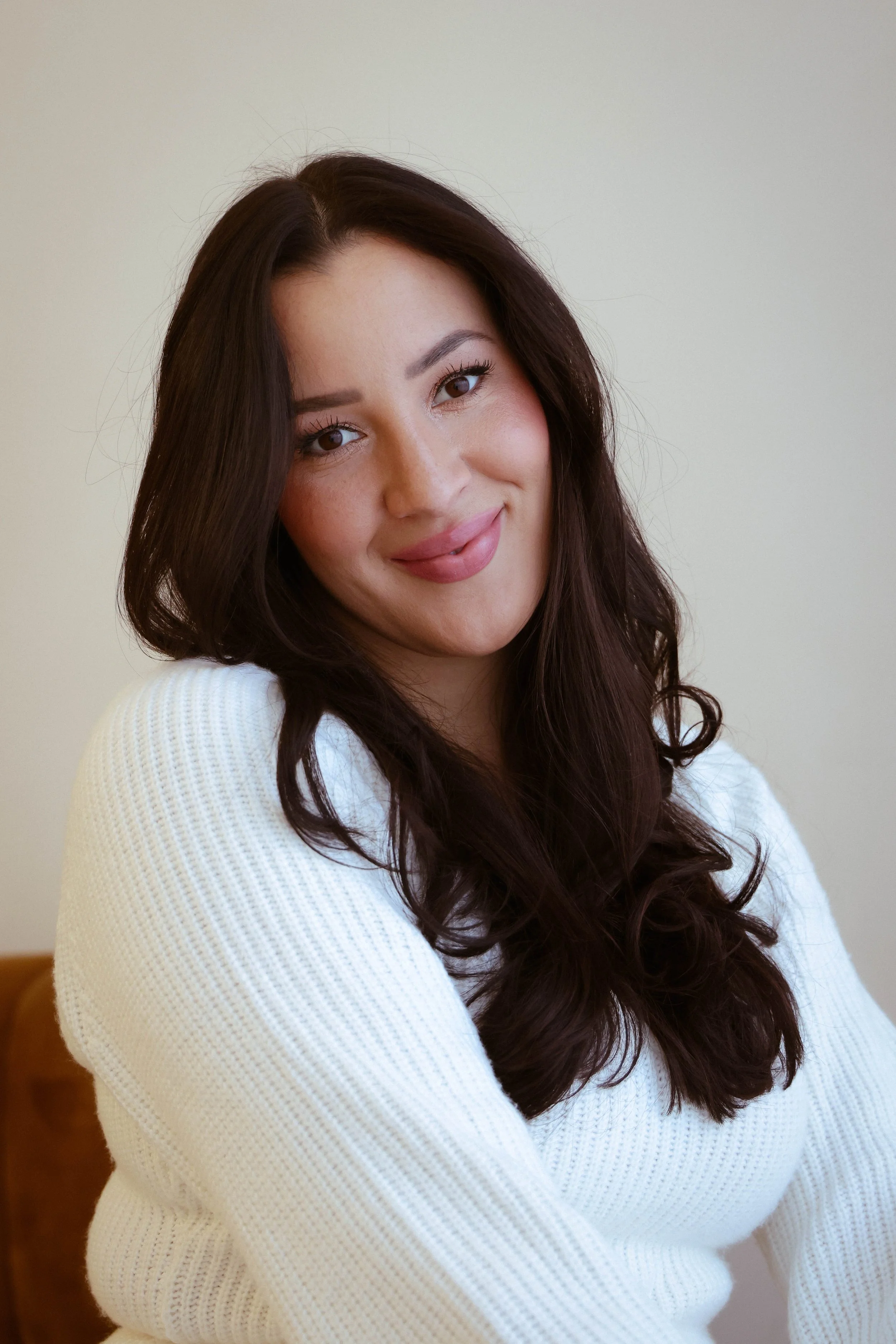 A woman with long, dark brown hair wearing a white sweater, smiling and sitting on a chair against a plain light-colored background.