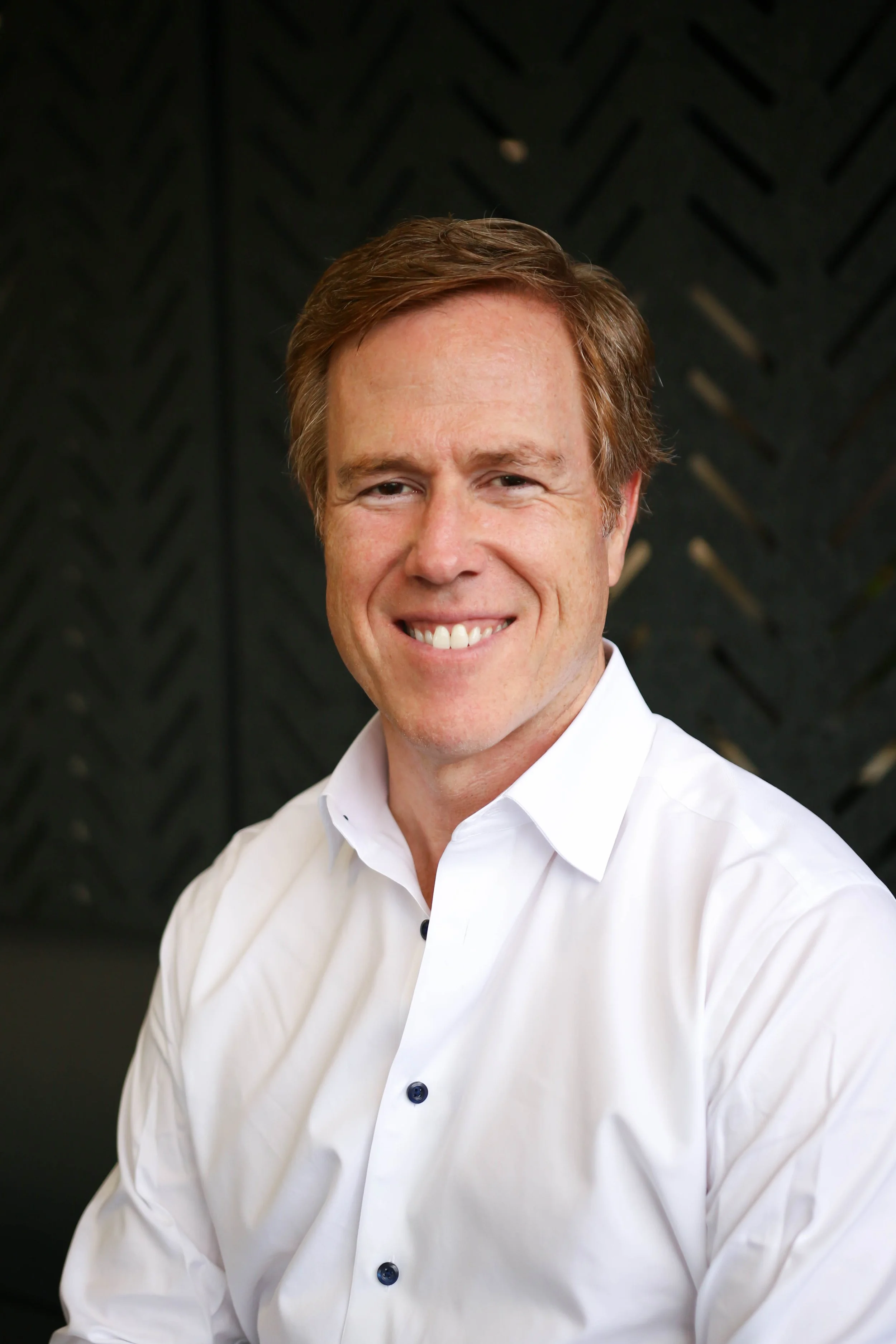 A smiling man with reddish-brown hair wearing a white button-up shirt, sitting against a dark background with a geometric pattern.
