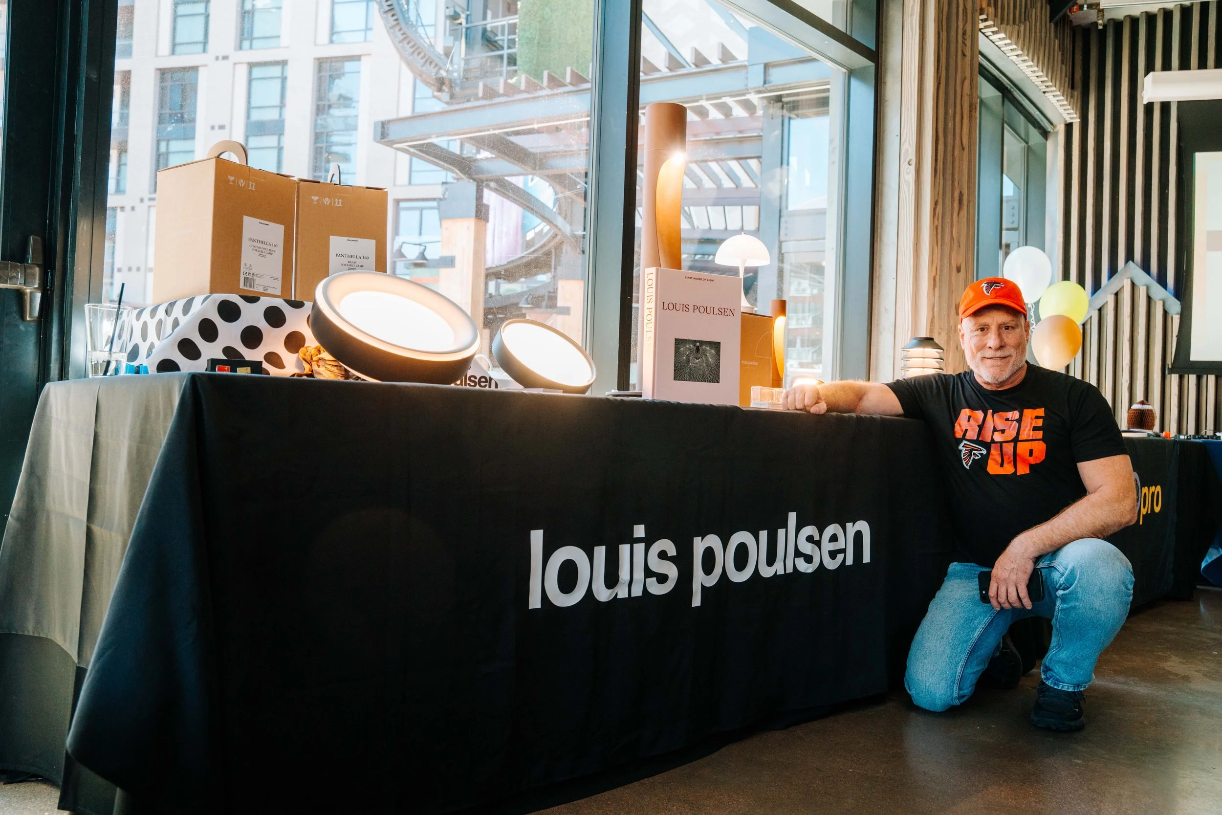 A man with a beard wearing a red cap and black T-shirt sitting on the floor next to a table covered with a black tablecloth that has "louis poulsen" written on it, with various lighting displays and boxes behind him in a modern indoor setting.
