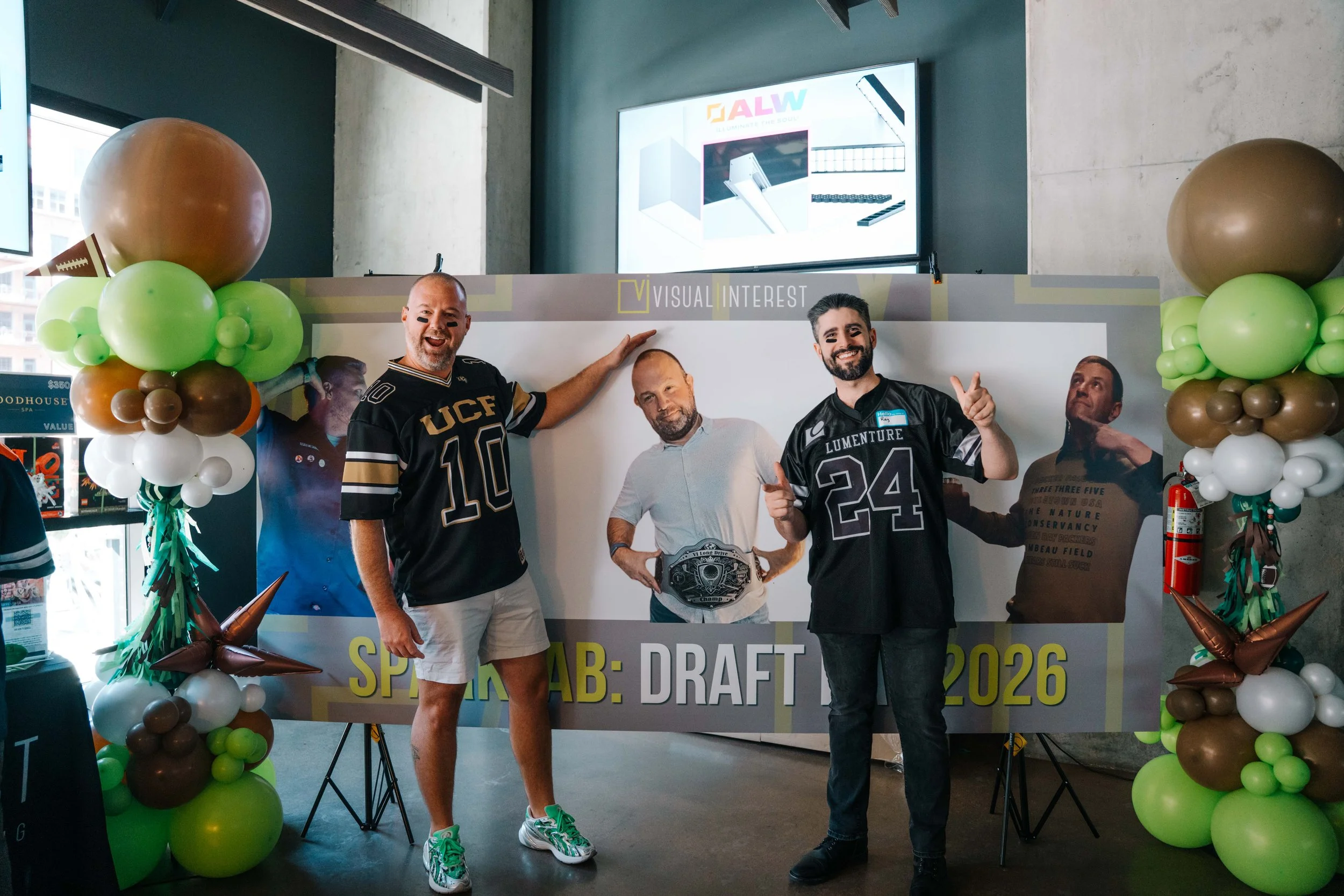 They are posing for a photo, smiling and making gestures. The man on the left is wearing a black sports jersey with the number 10, white shorts, and sneakers. The man on the right is wearing a similar sports jersey with the number 24, black pants, an