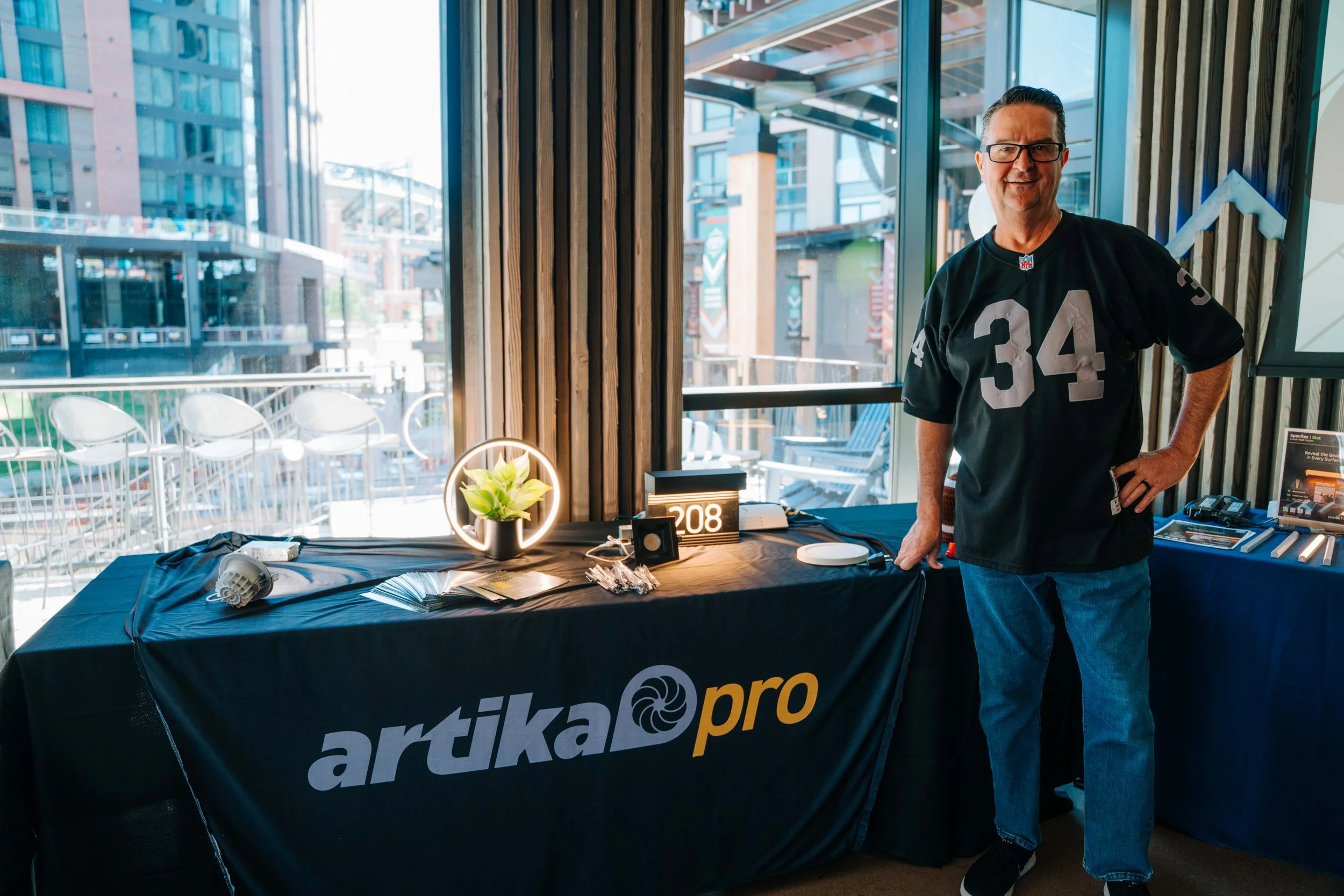 A man wearing glasses and a black football jersey with the number 34 stands behind a table with promotional materials and a sign reading 'artika pro'. The table has a black tablecloth with the 'artika pro' logo, and there is a small plant and a clock
