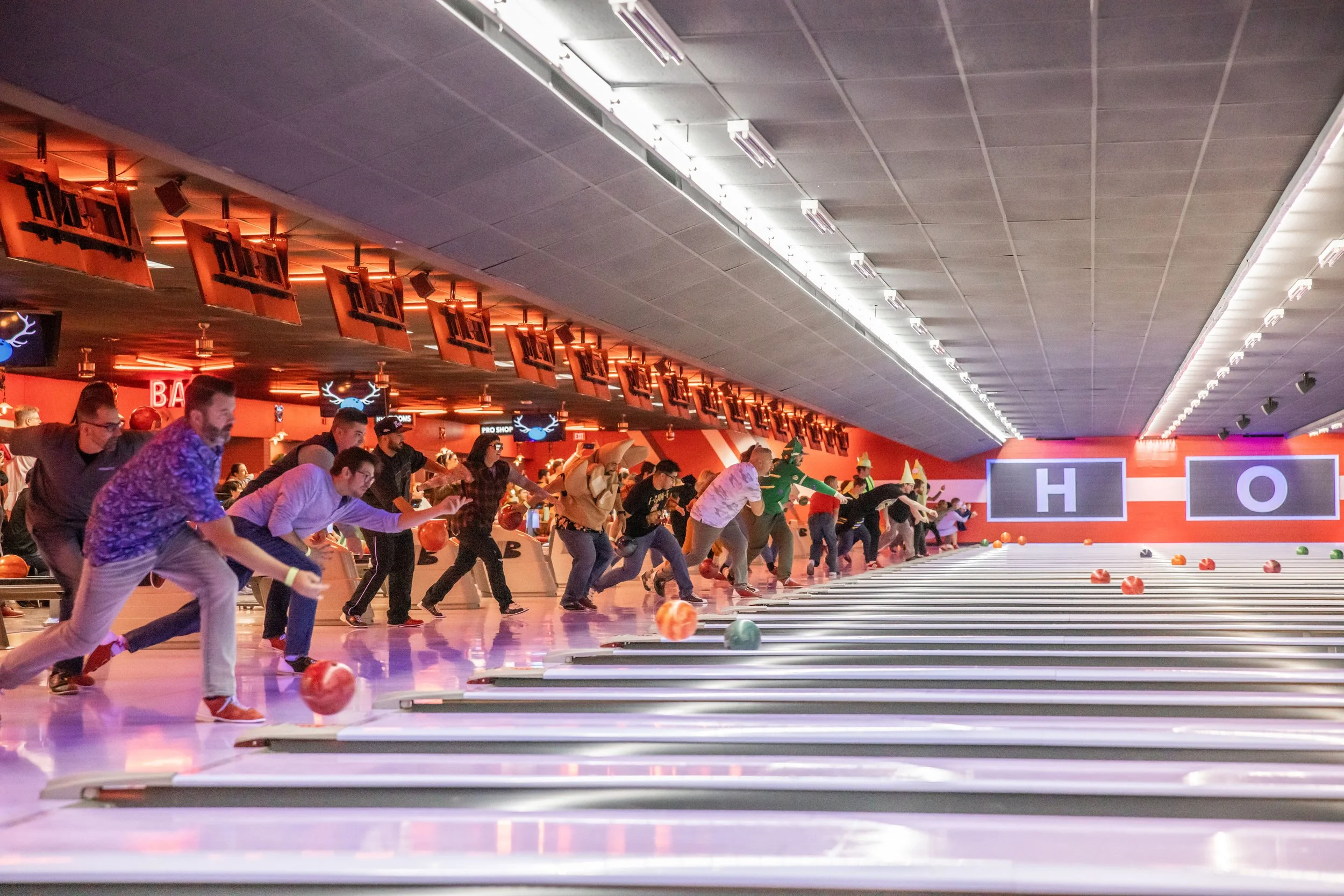 People bowling in an indoor alley, some wearing costumes, with screens displaying letters 'H' and 'O' at the end of the lane, and scattered bowling balls on the floor.