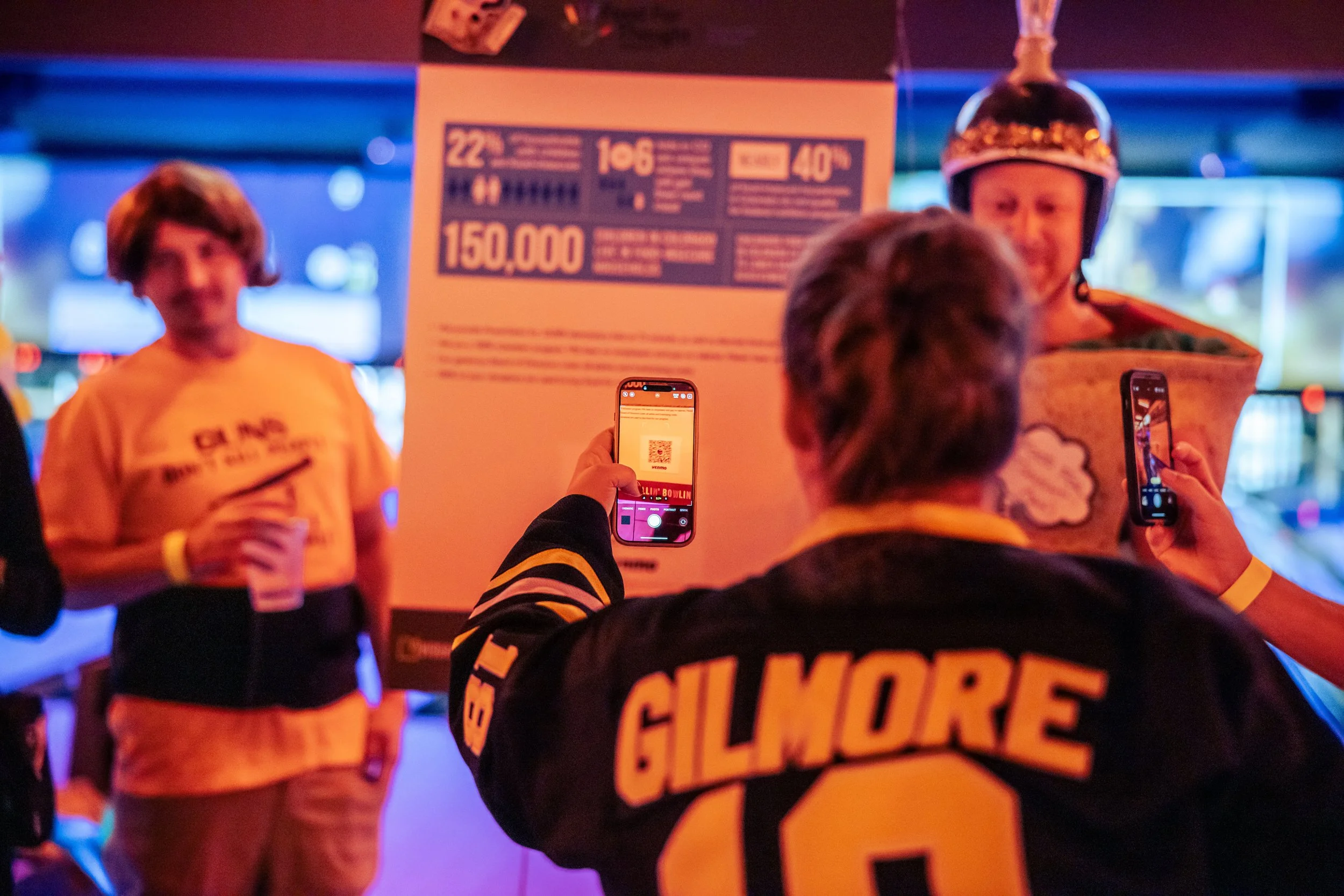 People at a sports event taking photos of a person in a costume around a poster with statistics, some holding drinks, with a person wearing a 'Gilmore' football jersey in the foreground.